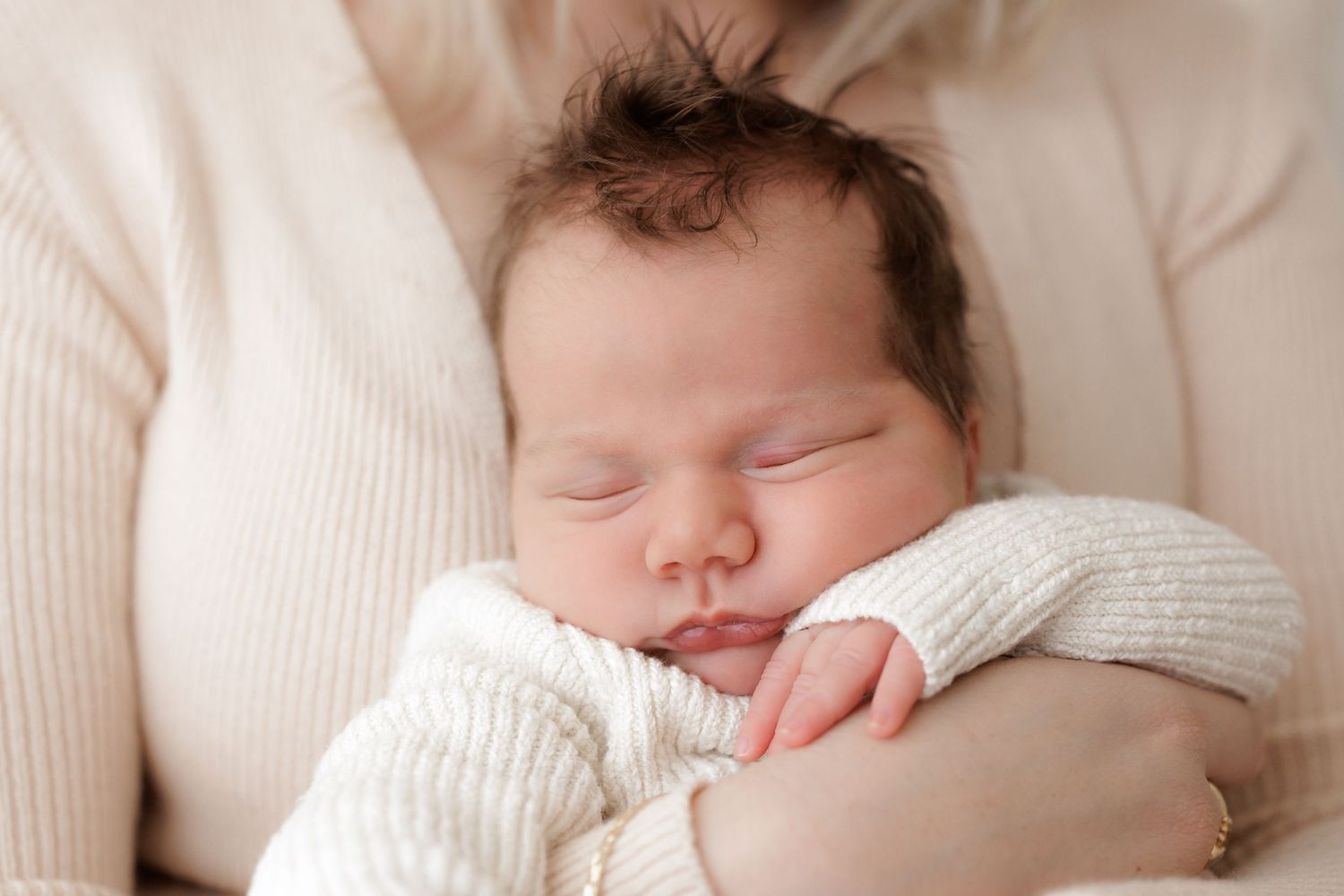 Sleeping newborn with spiky dark hair wearing white knit sweater.