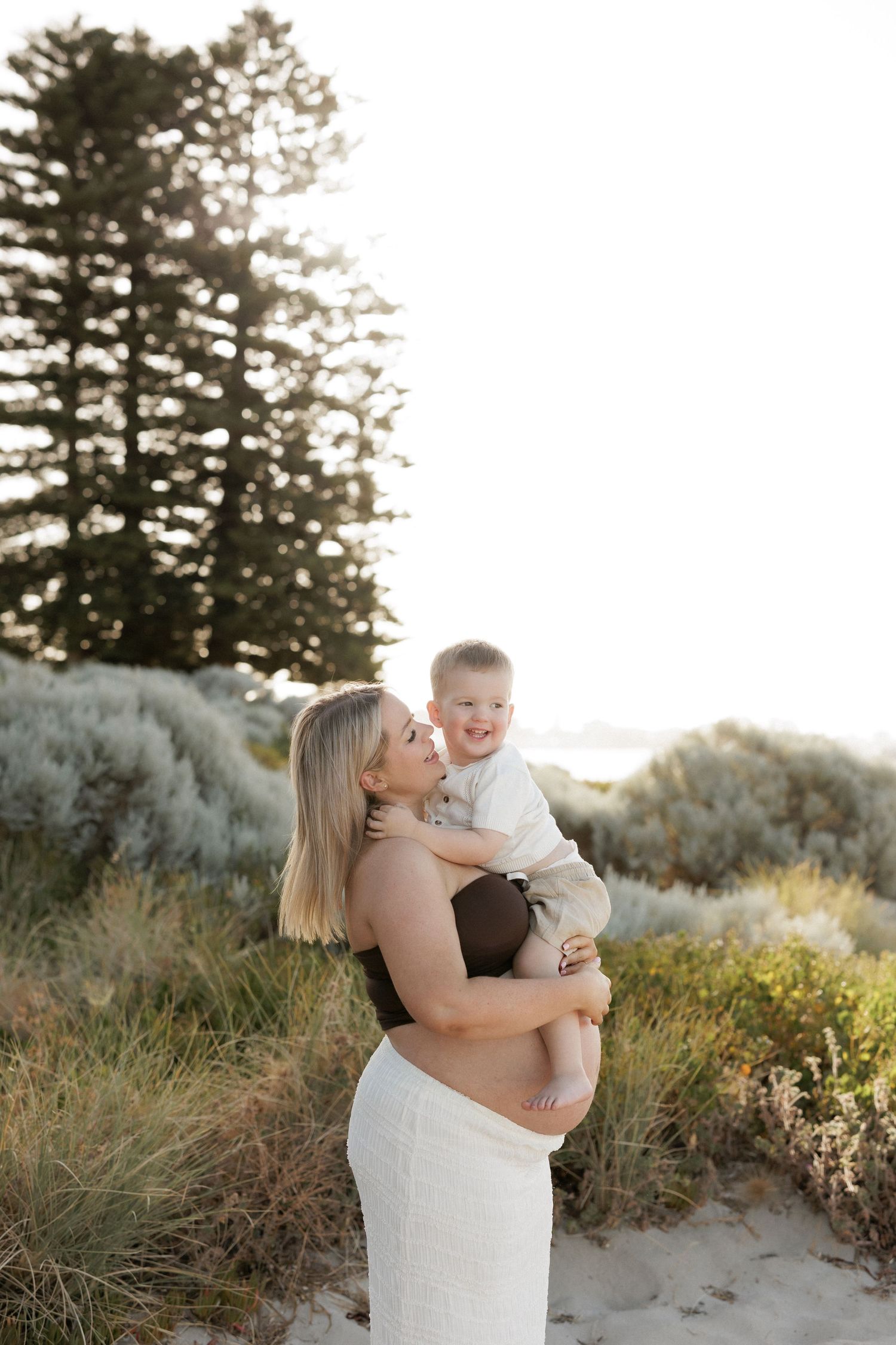 Pregnant woman in white skirt holding smiling baby outdoors near beach with green shrubs and pine trees in background.