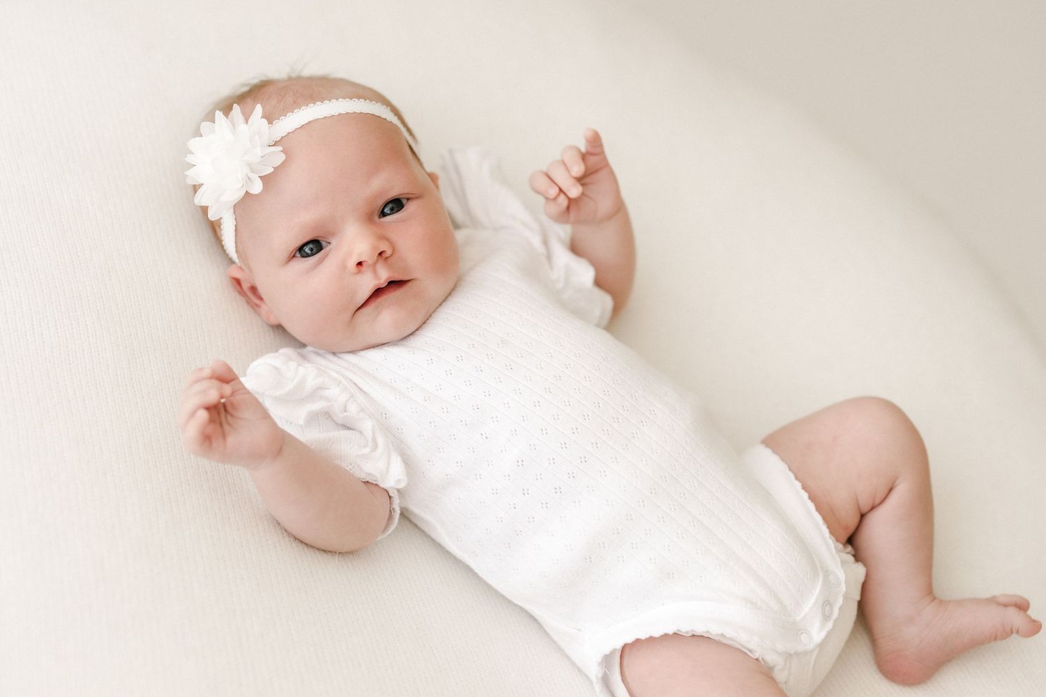 Baby in white romper with flower headband lying on cream backdrop.