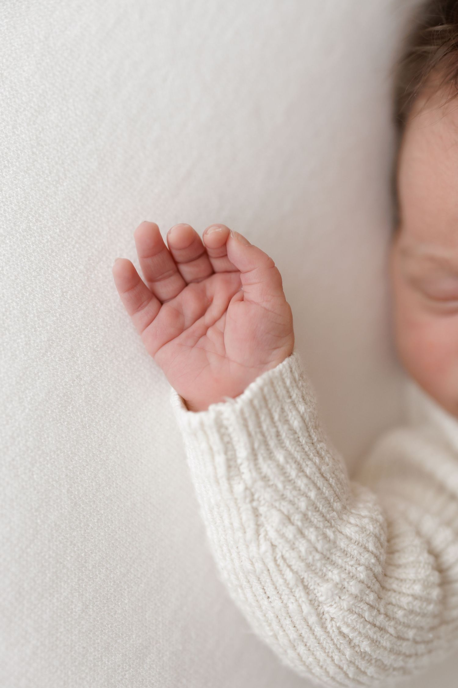 Close-up of tiny baby hand wearing white knit sweater against light background.