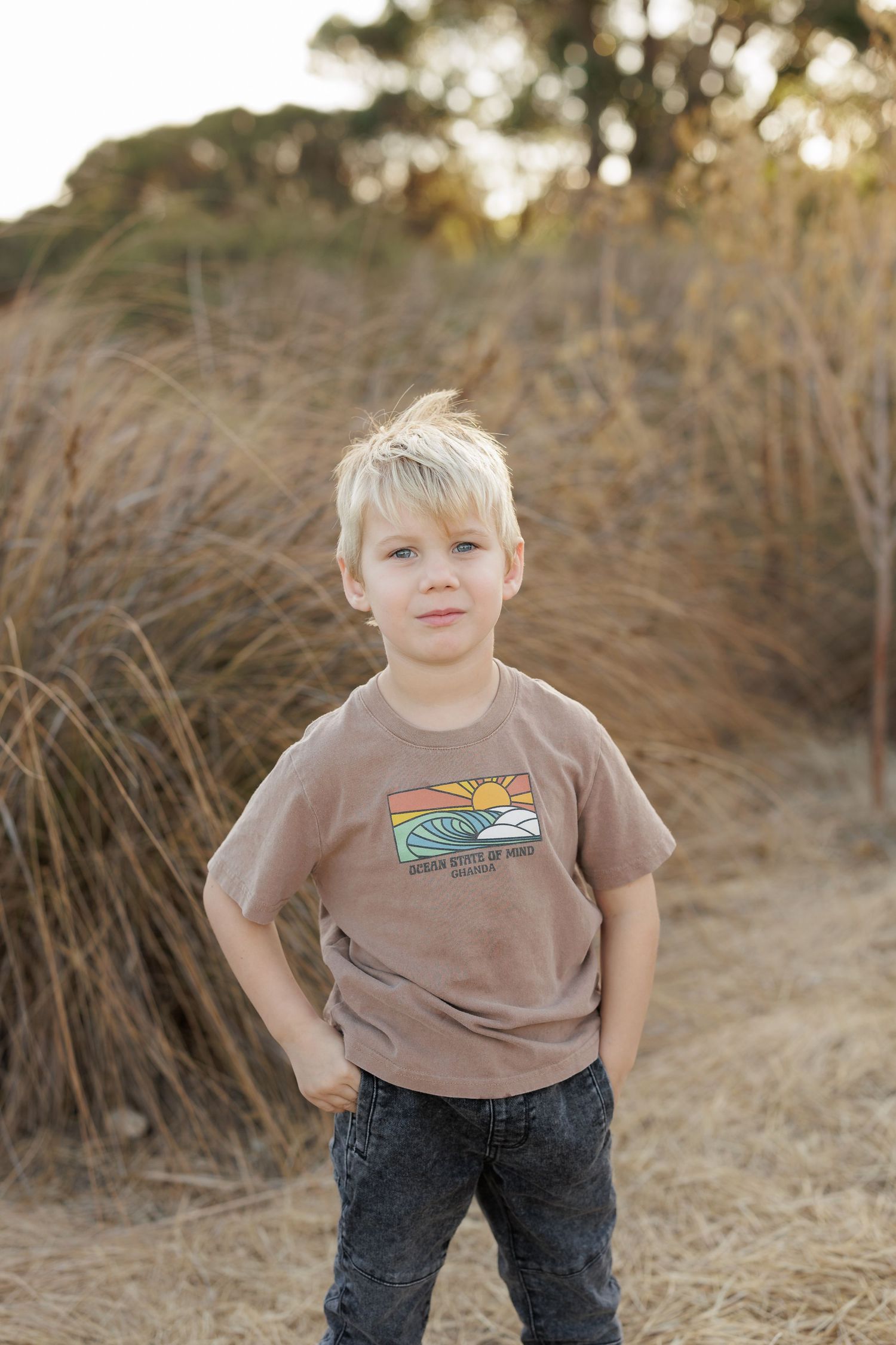Young boy in brown graphic t-shirt poses outdoors among tall grass.