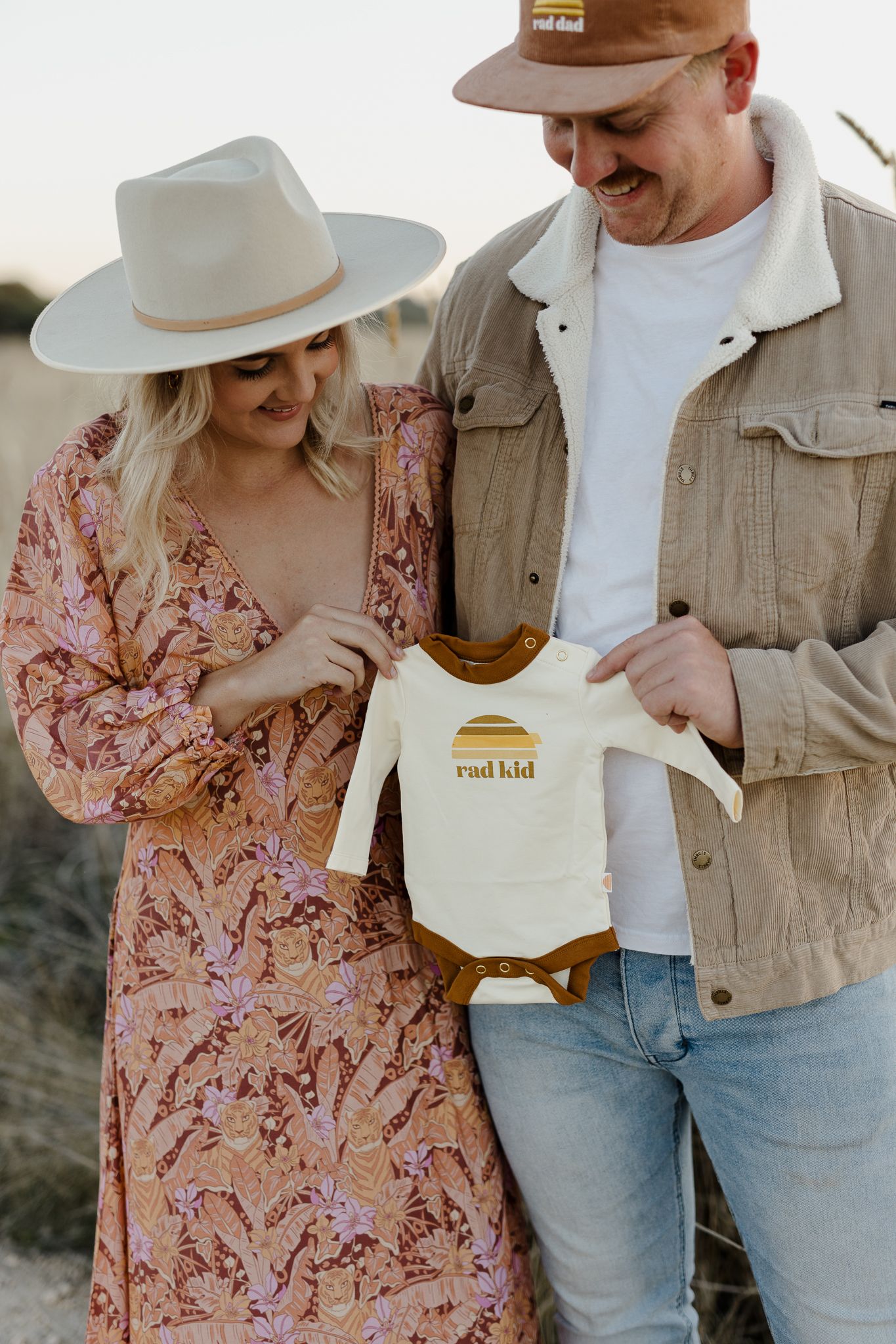 Couple holding up a small white onesie with text in a field at sunset.