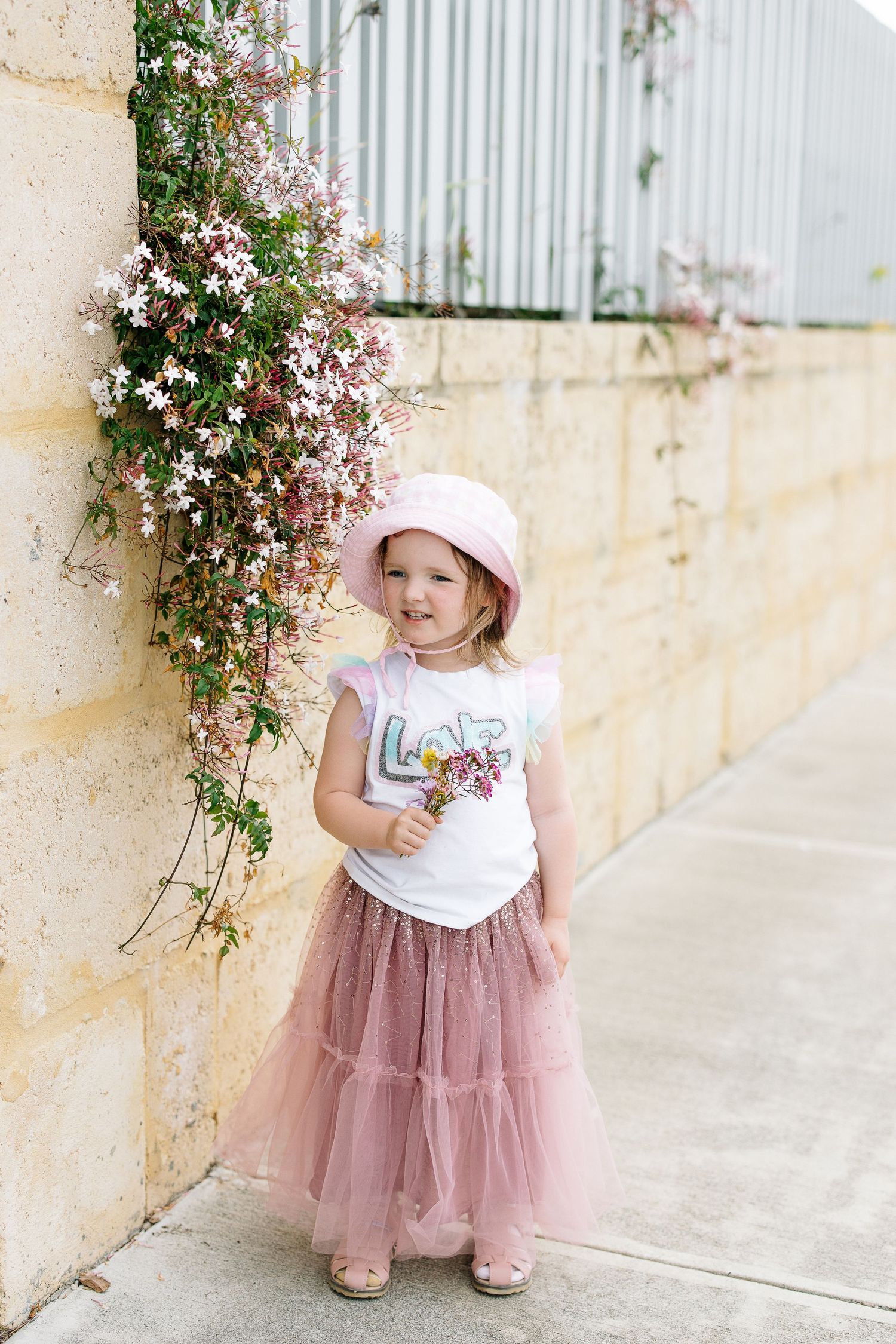 A delicate pink tulle skirt and sun hat outfit photographed against a wall with climbing flowers.