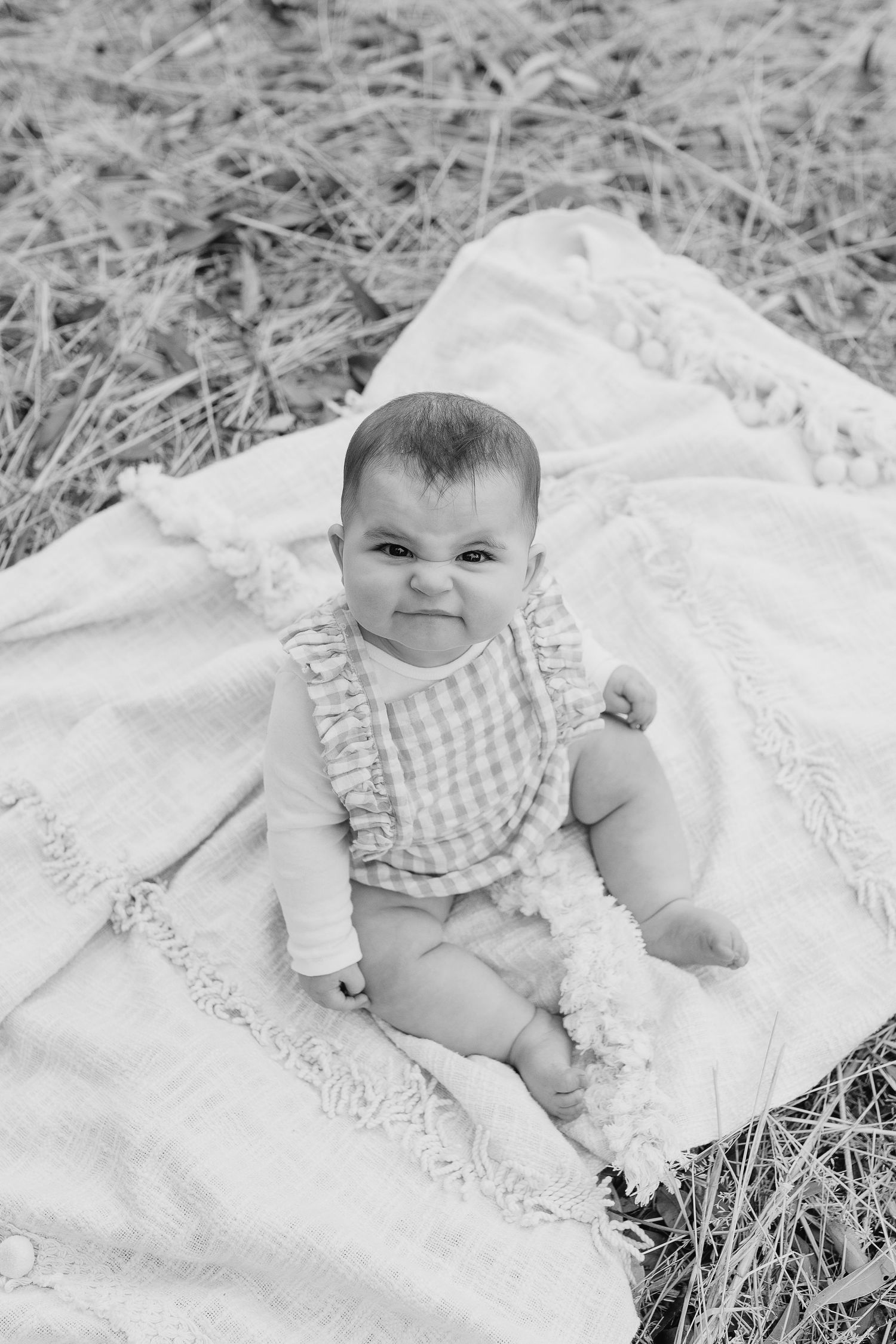 Black and white portrait of baby in ruffled outfit sitting on white blanket outdoors.