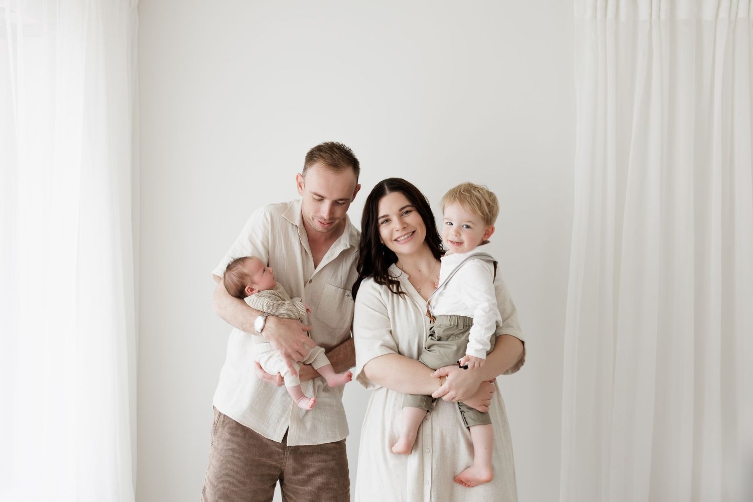 Family wearing neutral colors poses together against white curtain backdrop in studio setting.