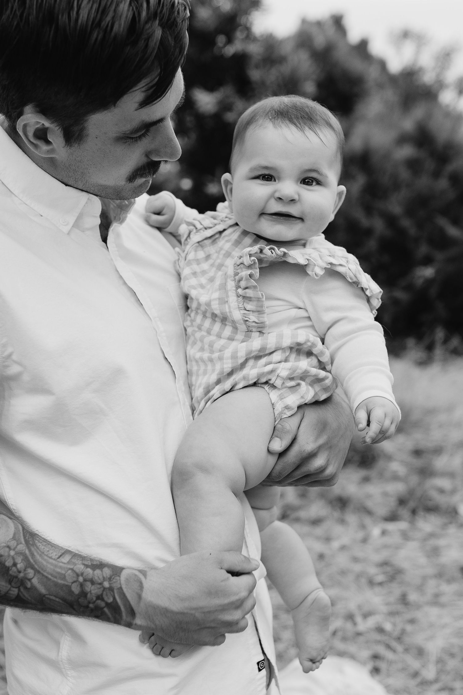 Black and white photo of parent holding happy baby in gingham outfit outdoors.