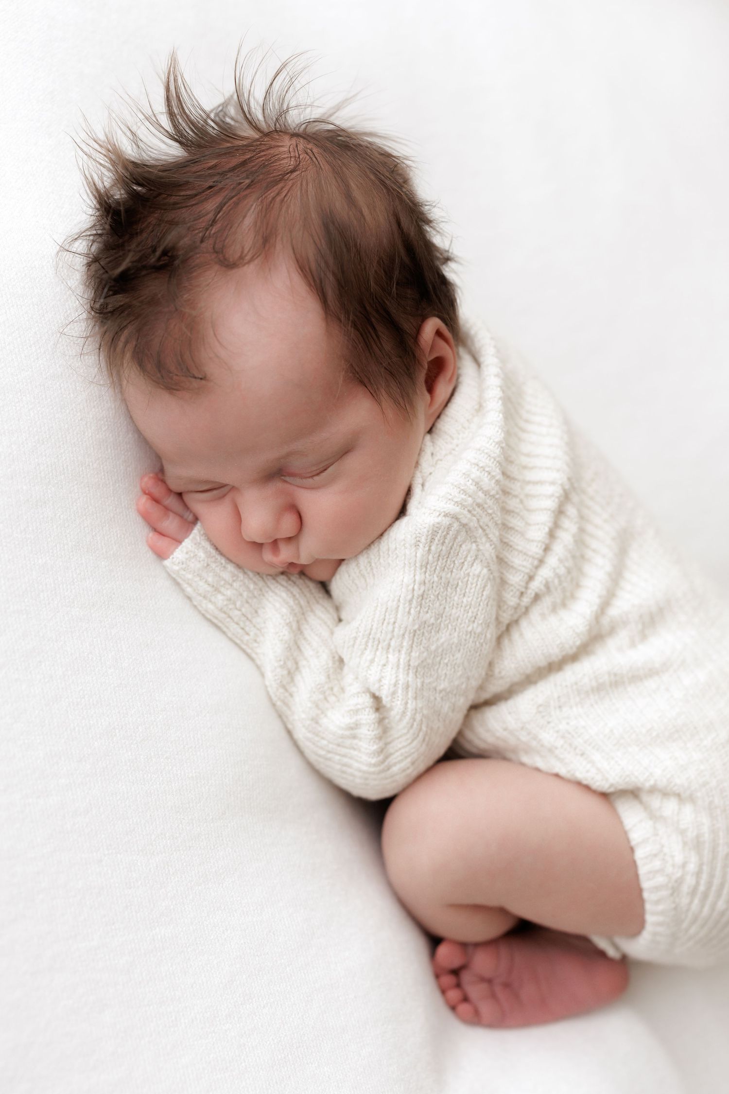 Sleeping infant in white sweater curled up on soft white surface.