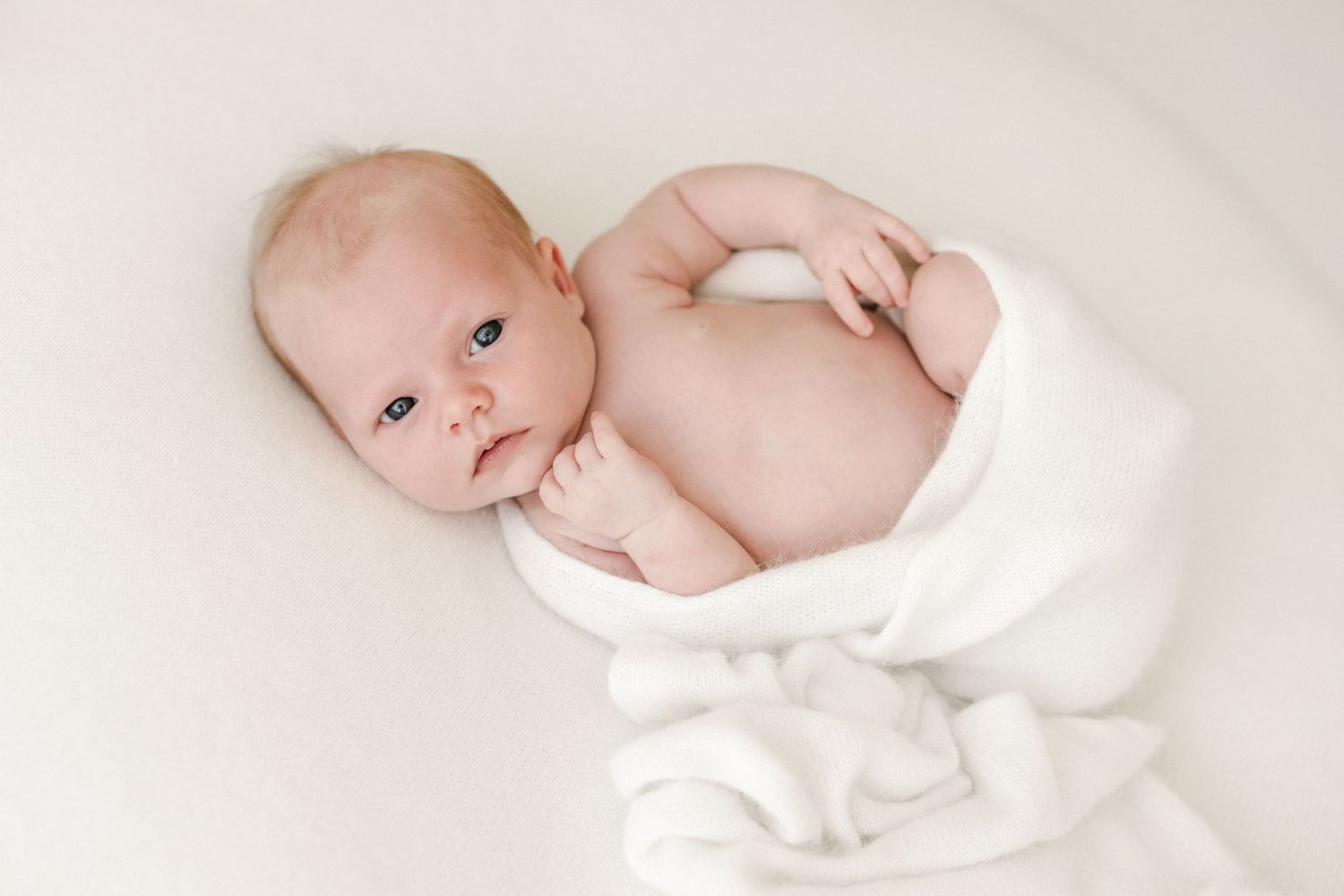 Newborn baby wrapped in white blanket on cream colored background.