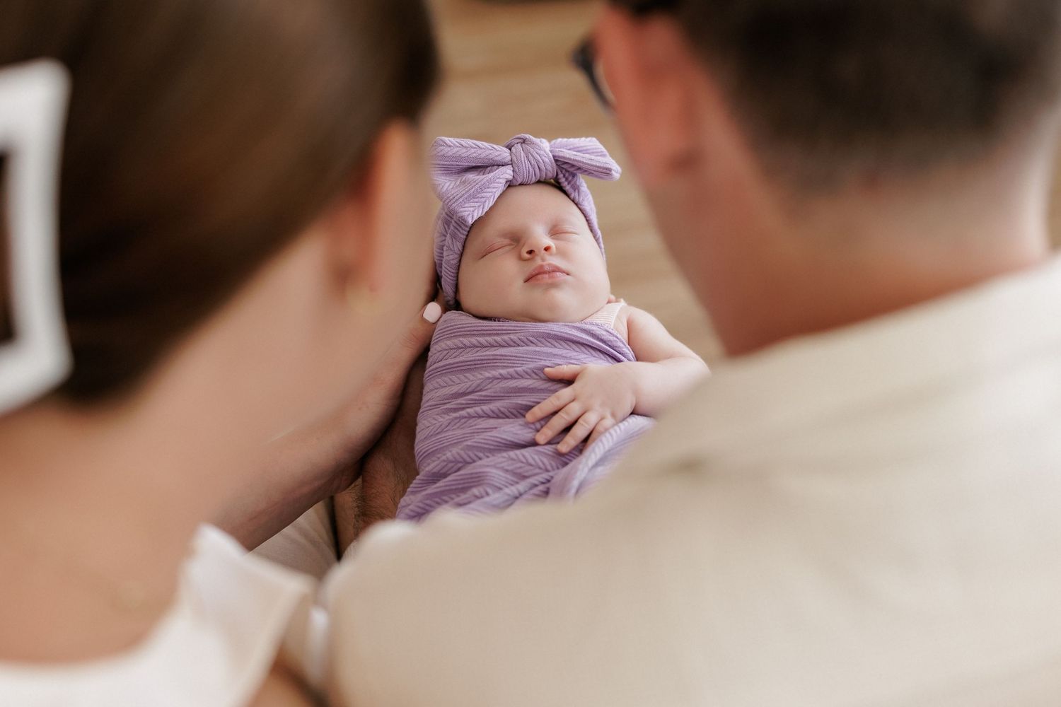 Newborn baby swaddled in purple blanket with bow headband, cradled by parents in a tender family moment.