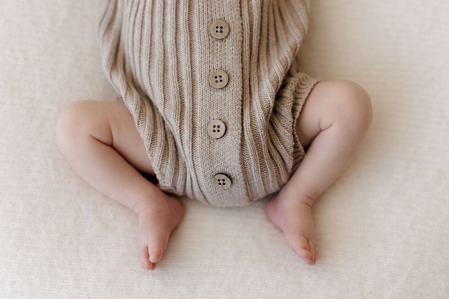 Close-up of a baby in a beige knit button romper, showing chubby legs and bare feet on a soft cream blanket.