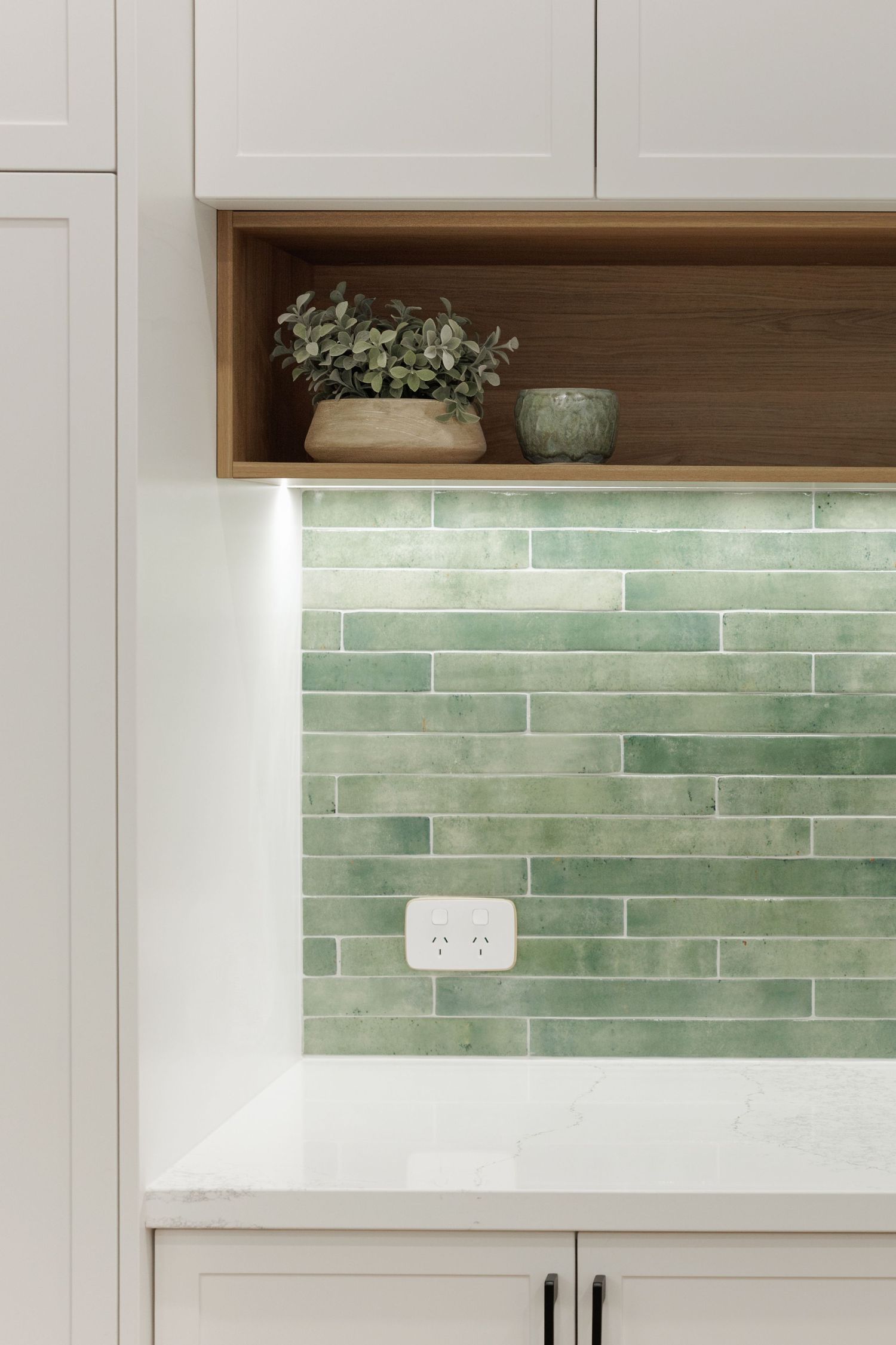 Modern kitchen backsplash with green subway tiles, white cabinets, marble countertop, and wooden shelf with plants.
