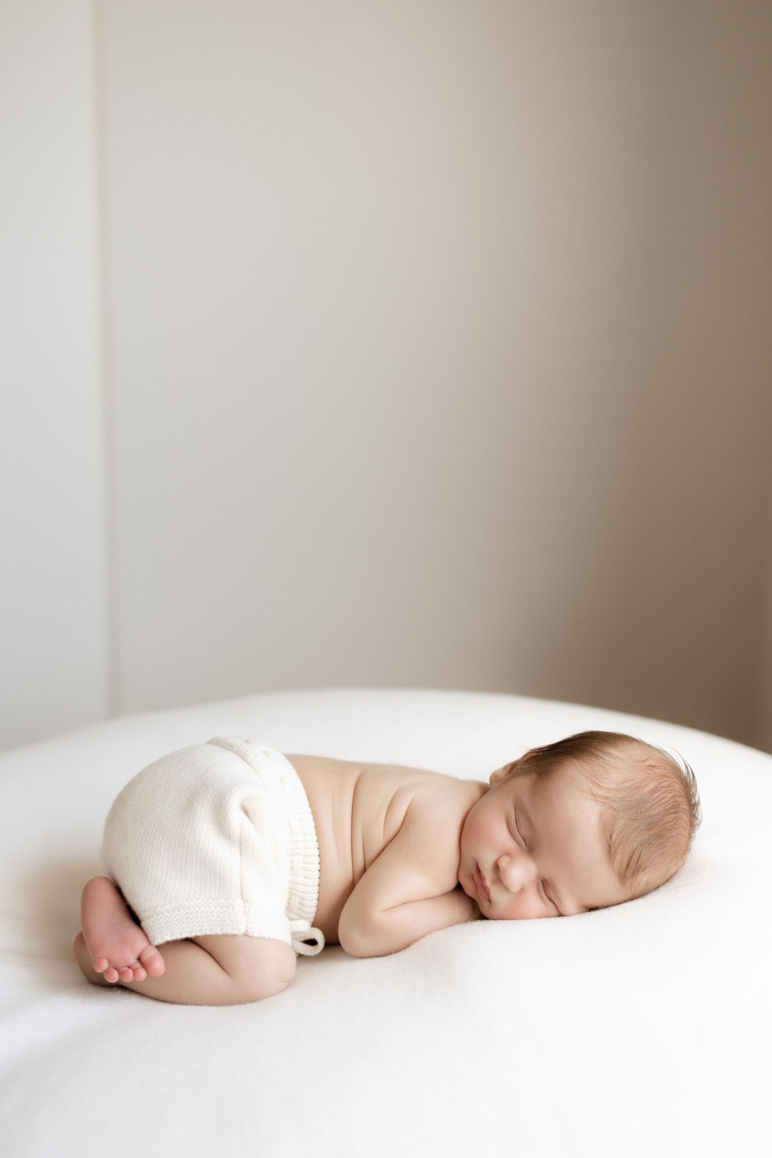 Sleeping newborn in white outfit curled up on soft white surface.