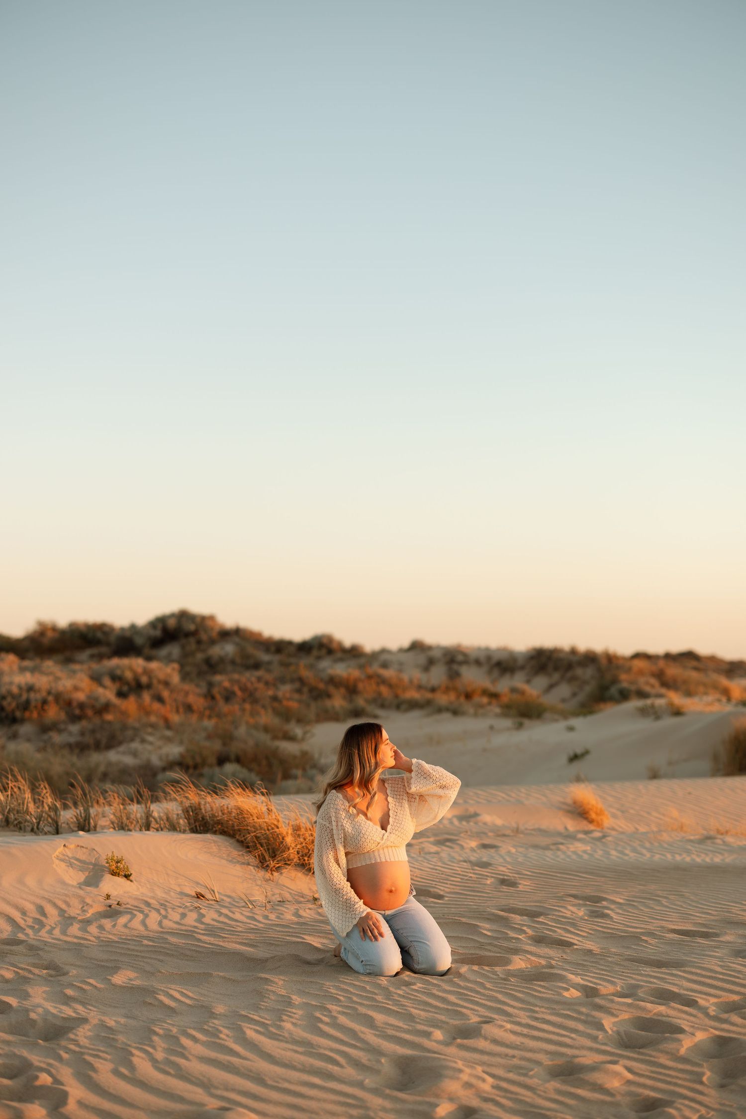 Person kneeling in light clothing on sandy dunes during sunset.