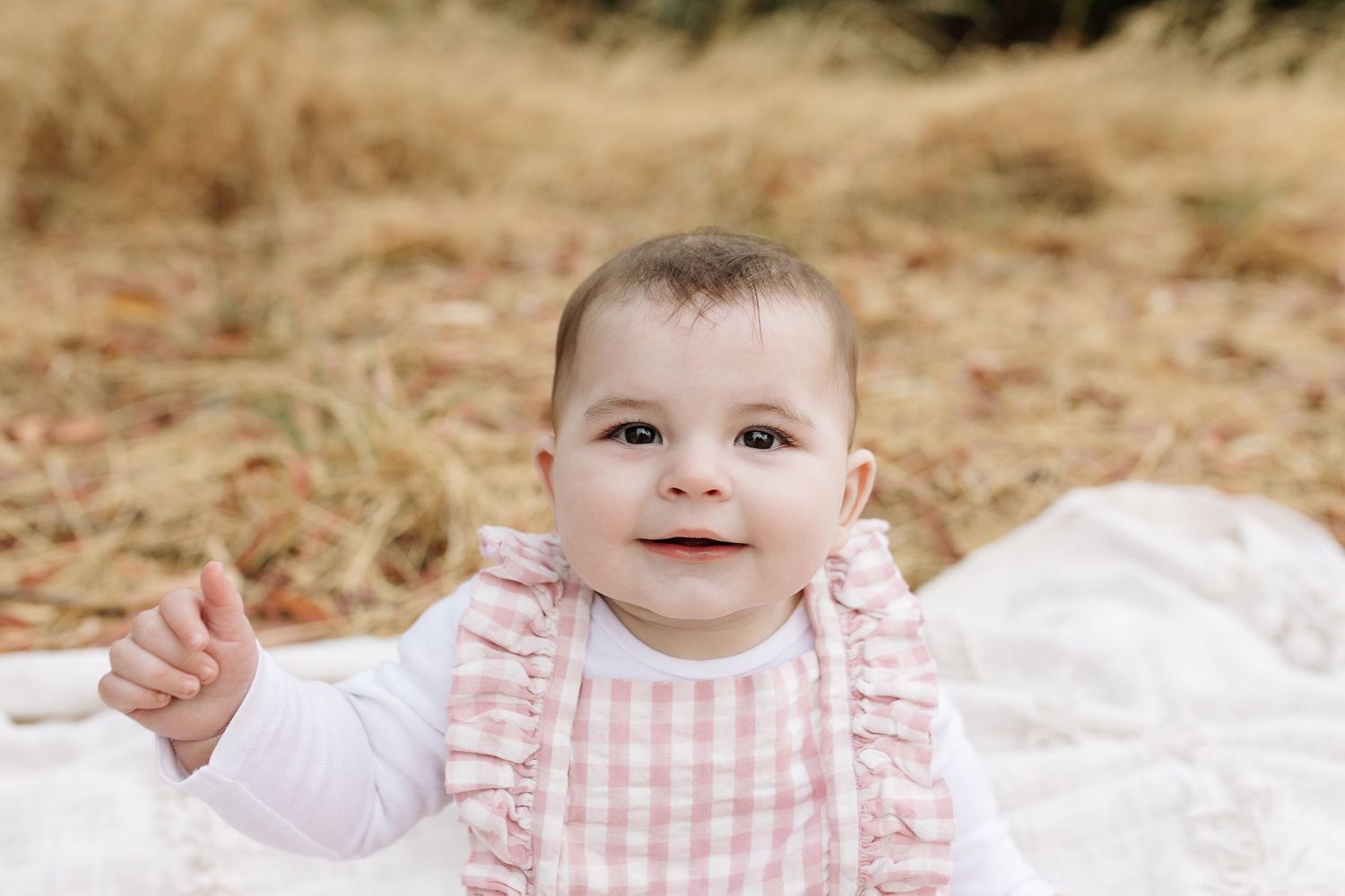 Happy baby in pink gingham outfit waves while sitting on white blanket outdoors.