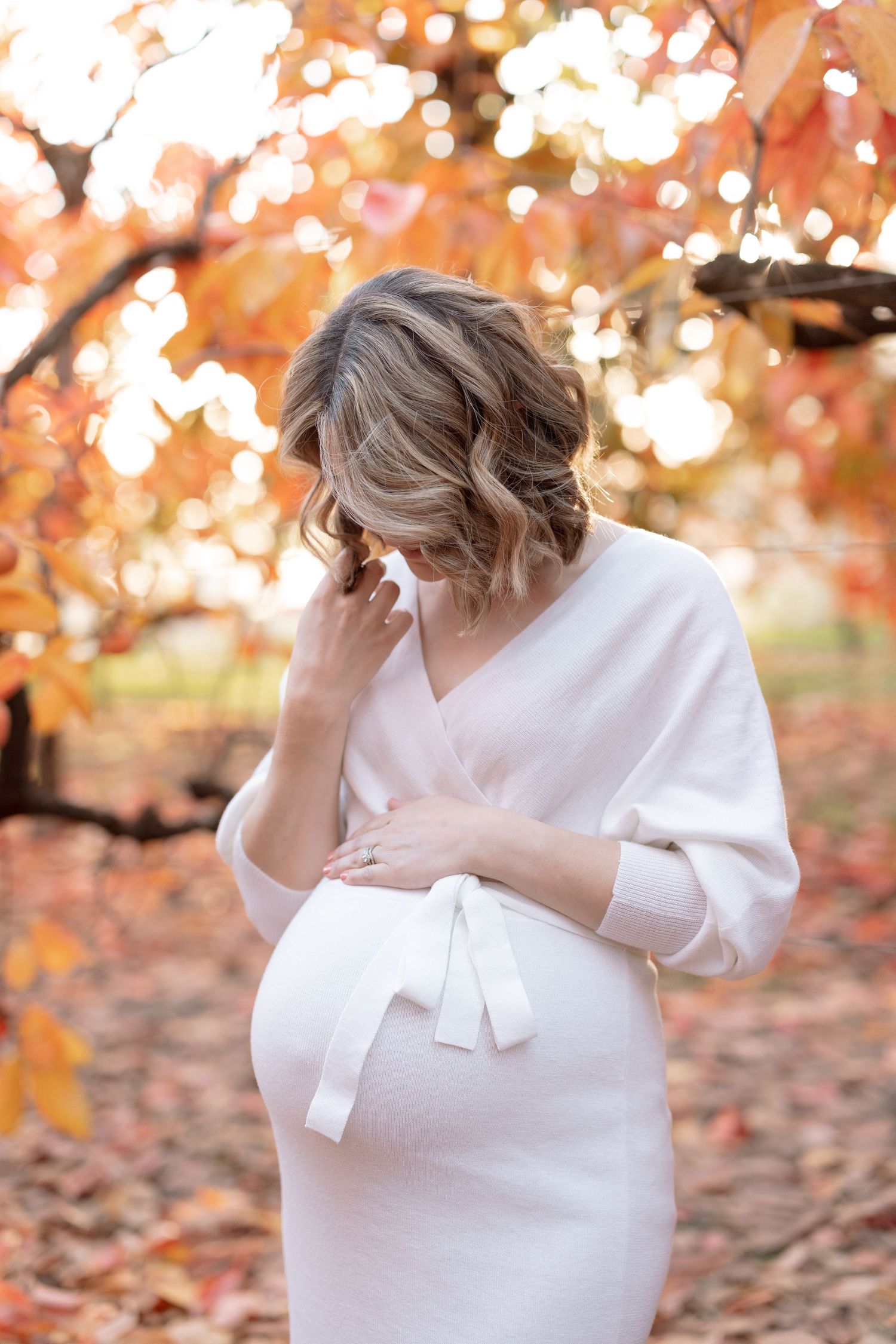 Maternity portrait showing woman in white dress under glowing fall foliage.