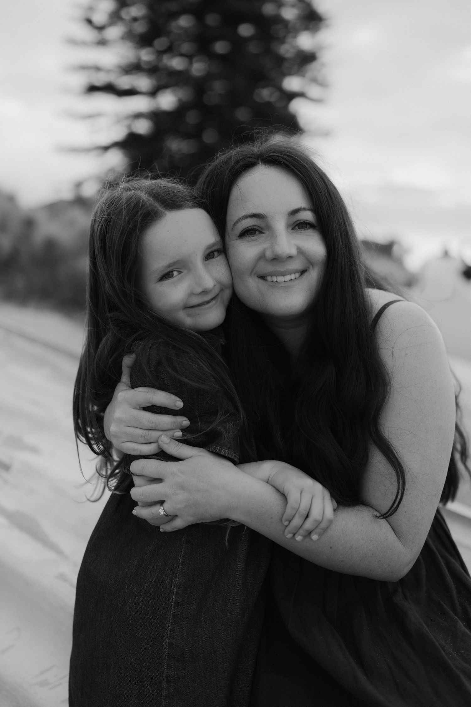 Black and white photo of a smiling mother and daughter hugging outdoors near a beach with a pine tree backdrop.