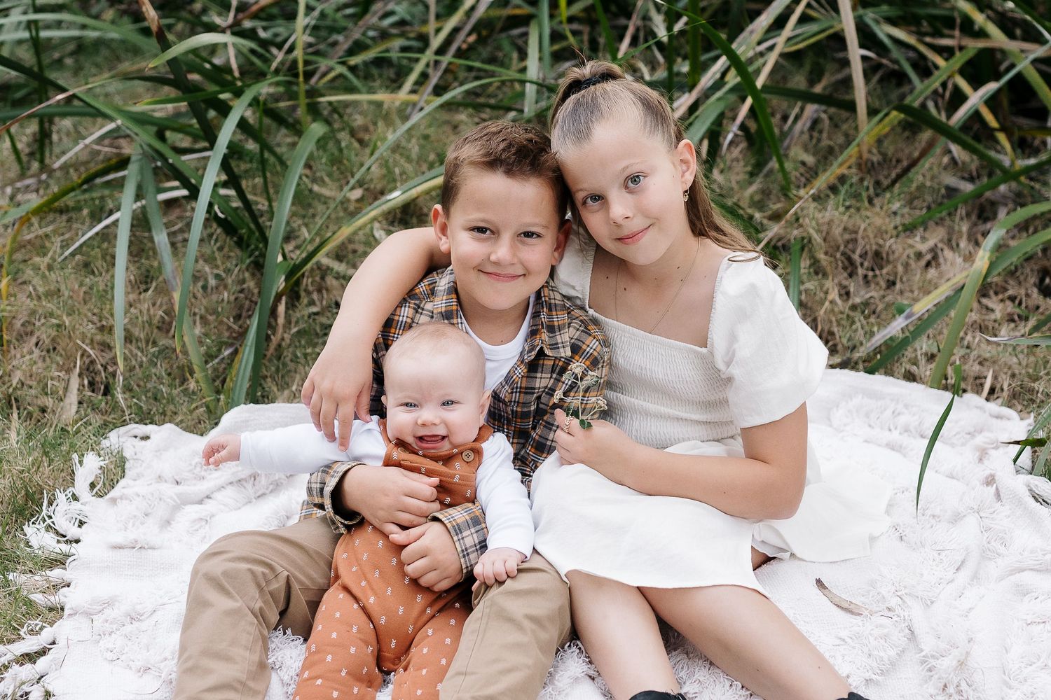 Three people sitting close together on white blanket surrounded by tall grass.