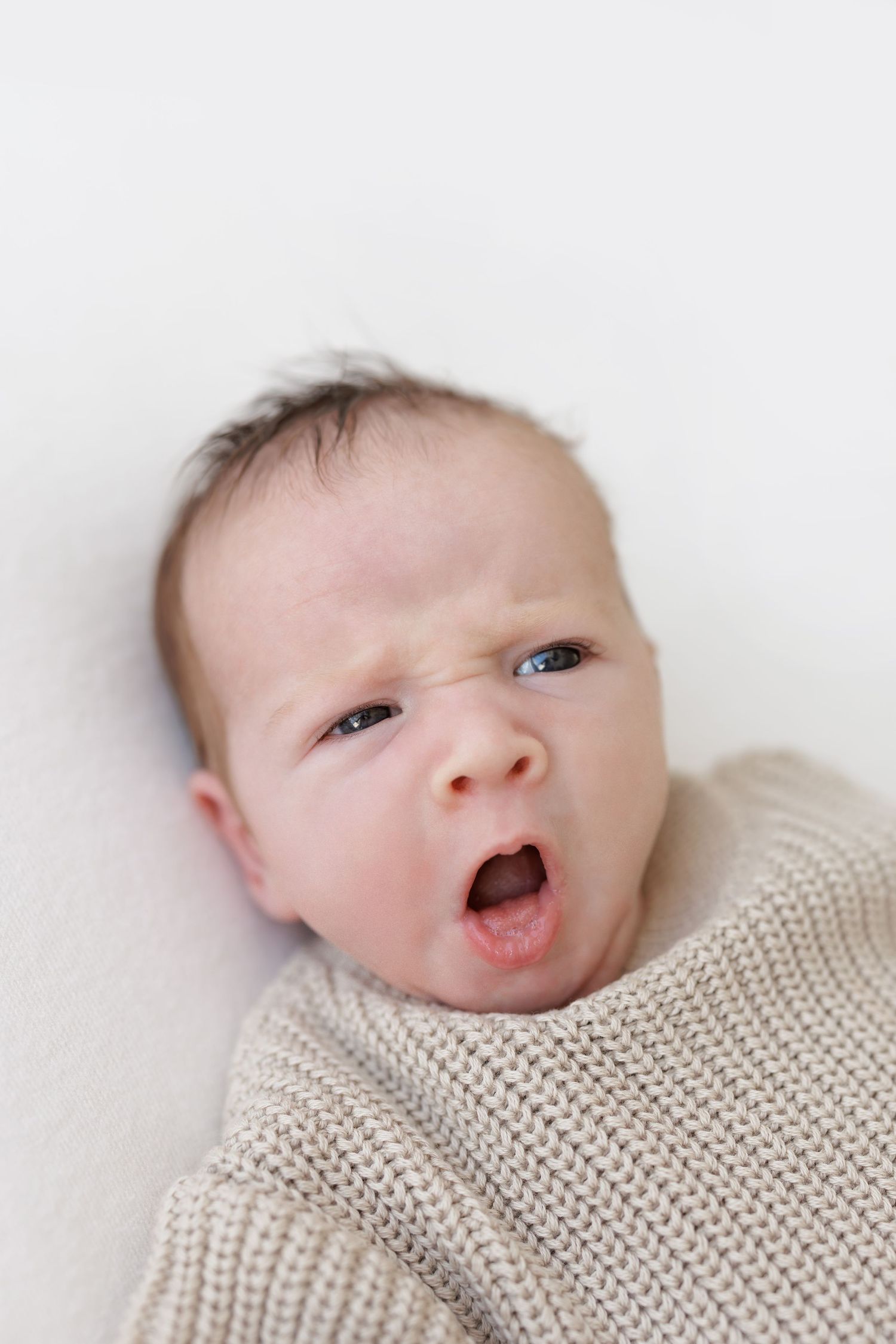 Baby wearing knit sweater yawns while lying on white background.