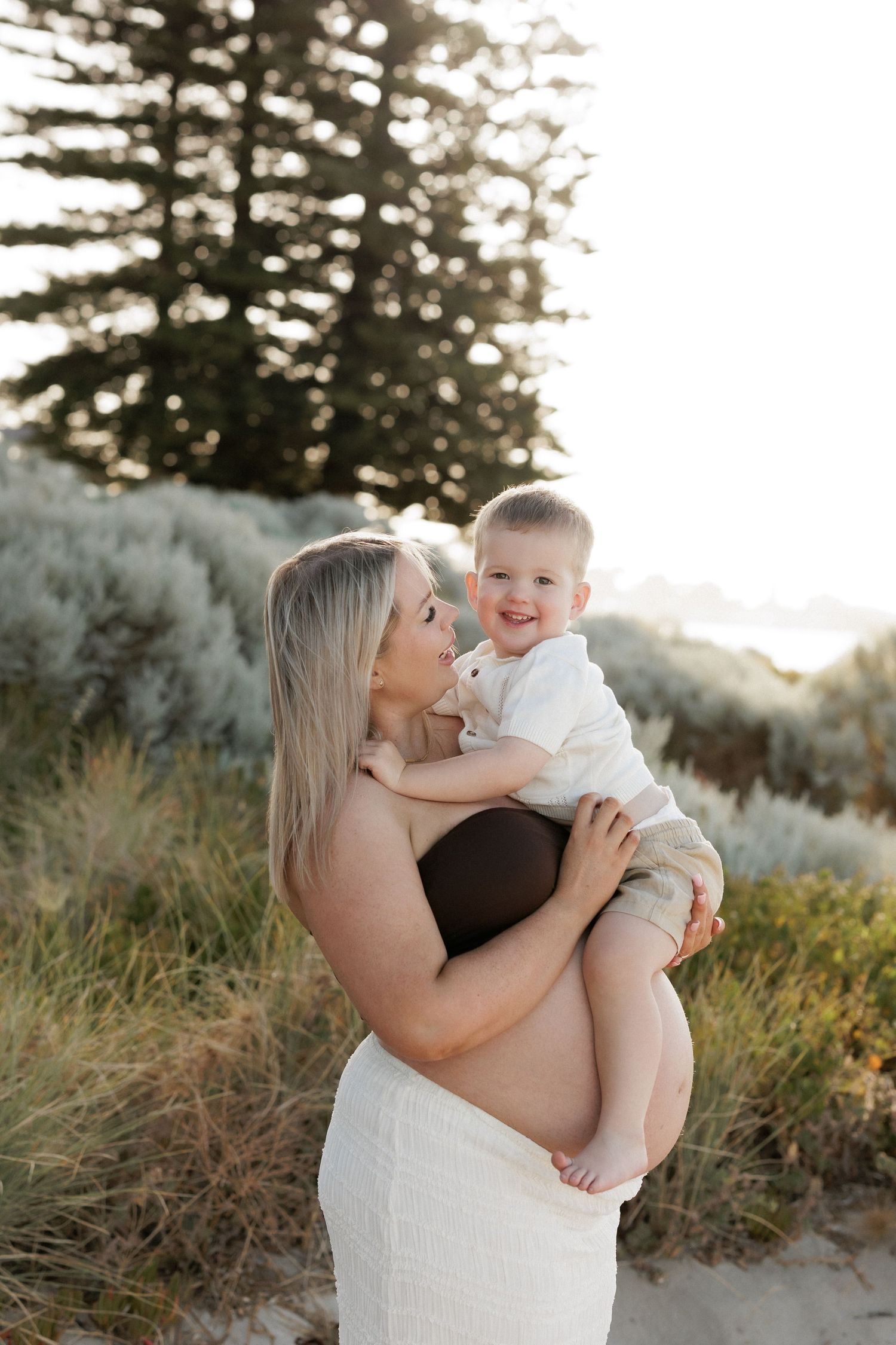 A tender outdoor maternity photoshoot in a natural setting with sunlit trees.
