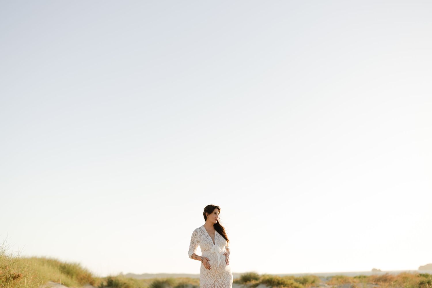 A woman in a white dress stands gracefully on a sandy beach with golden grass under a clear sky.