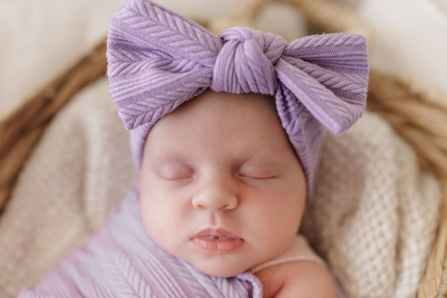 Sleeping newborn baby wrapped in purple swaddle wearing a lavender ribbed bow headband in a wicker basket.