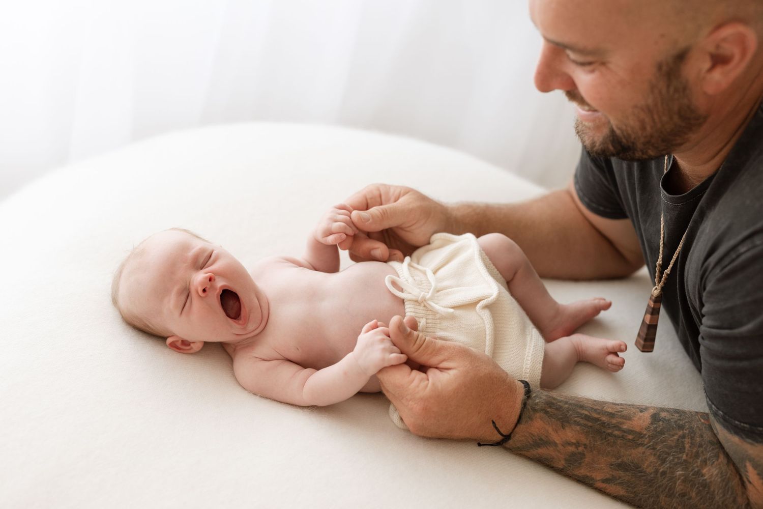 Man with tattoos playing with laughing baby on white surface during candid lifestyle photoshoot.