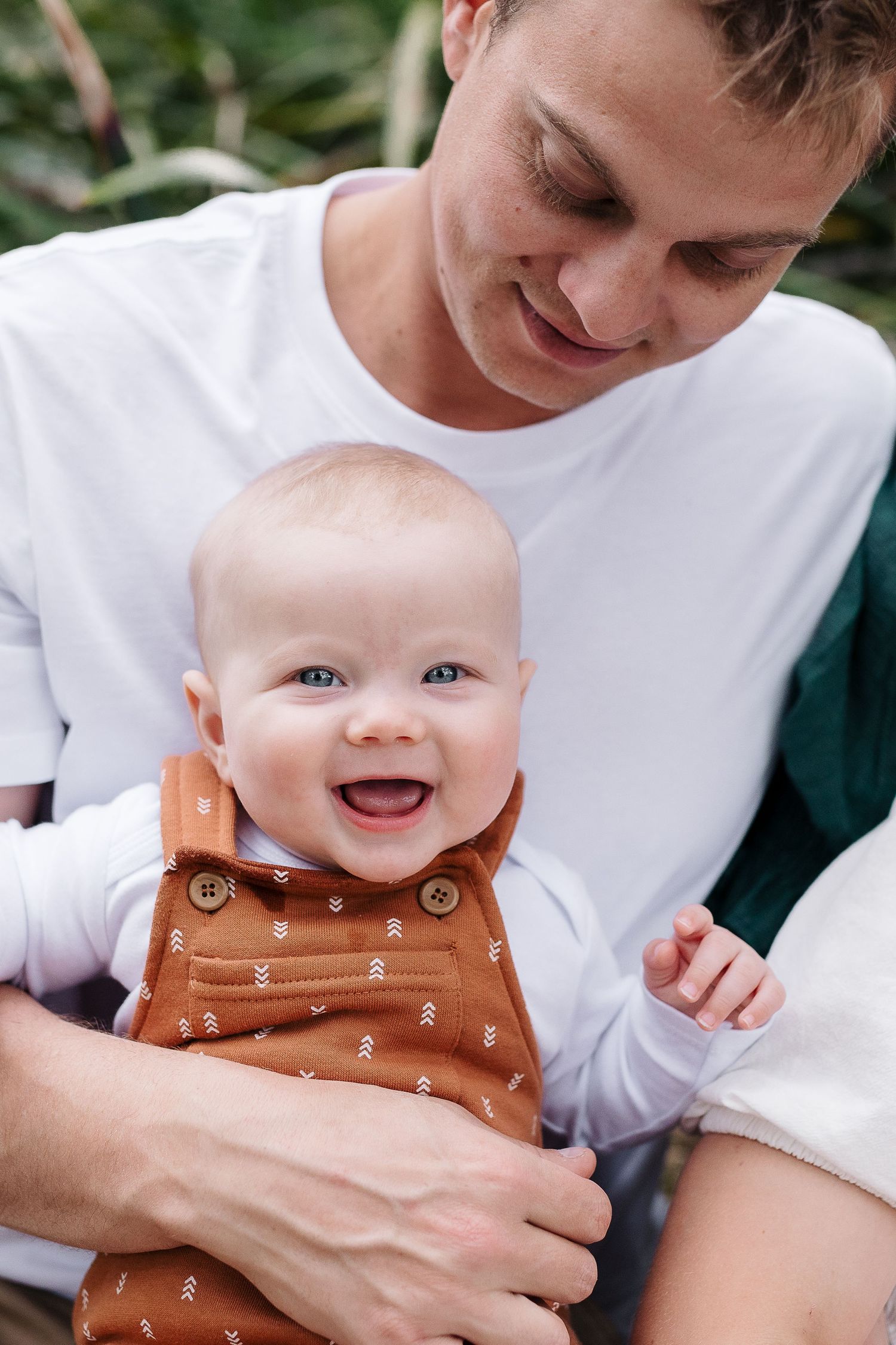 An adorable baby in brown overalls smiles while being held in an outdoor setting.
