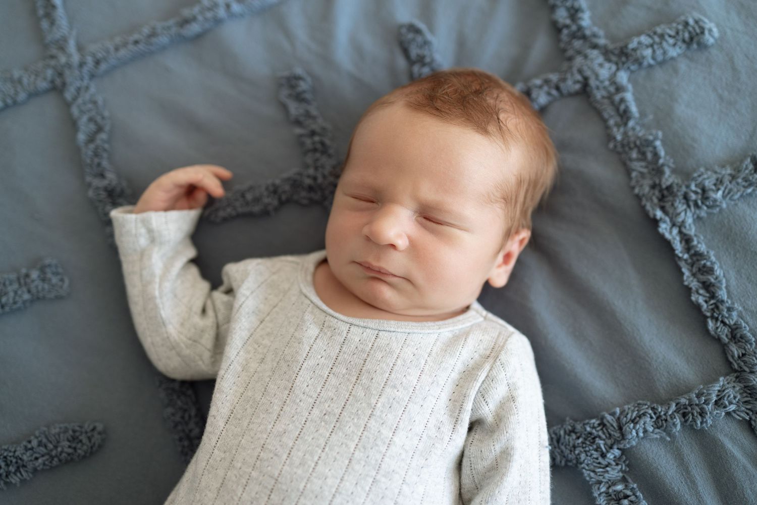 Peaceful newborn baby wearing cream sweater resting on textured gray blanket.