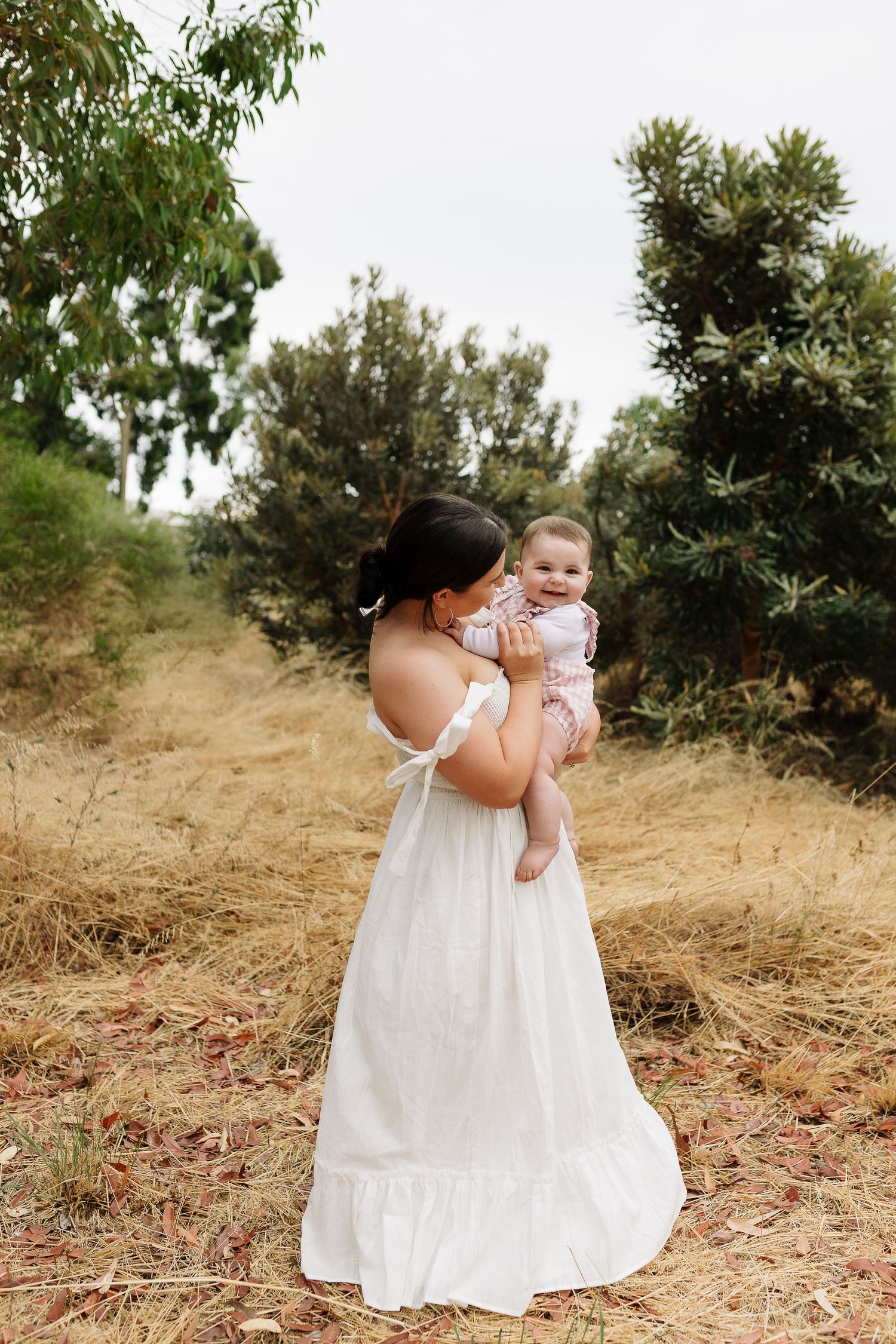 Person in white flowing dress holds baby against scenic natural backdrop with trees.