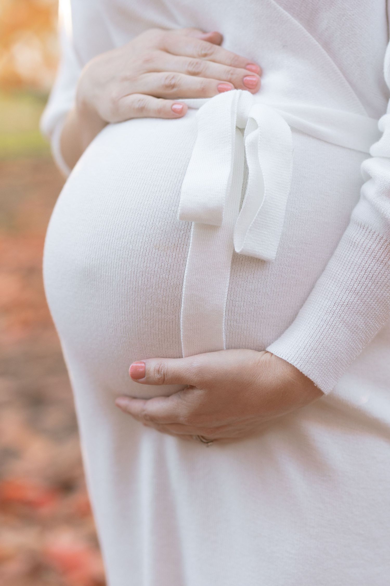Close-up of hands cradling pregnant belly in white dress with sash.