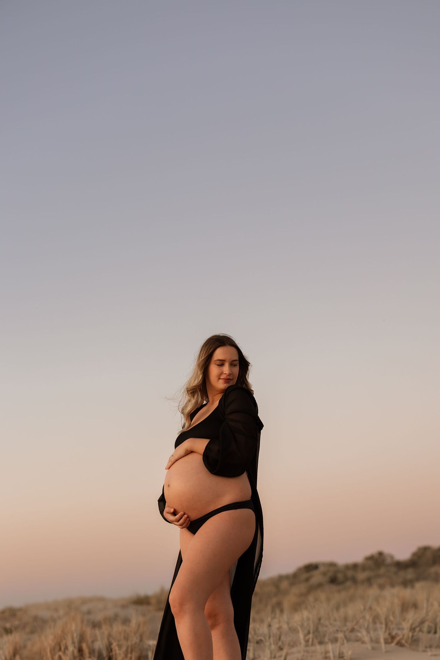 Silhouetted maternity portrait on sandy dunes at sunset showing a flowing black robe.