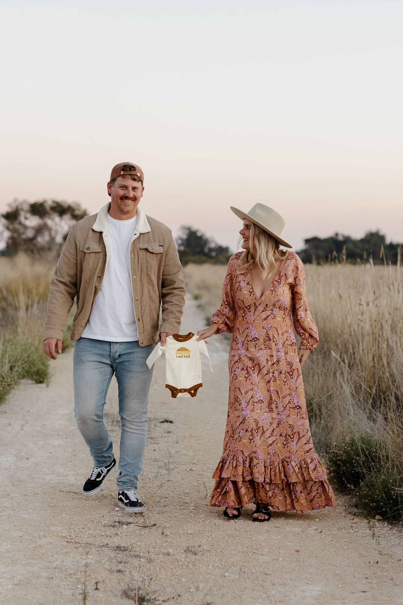 Couple walking on dirt path holding baby onesie between tall grass fields.