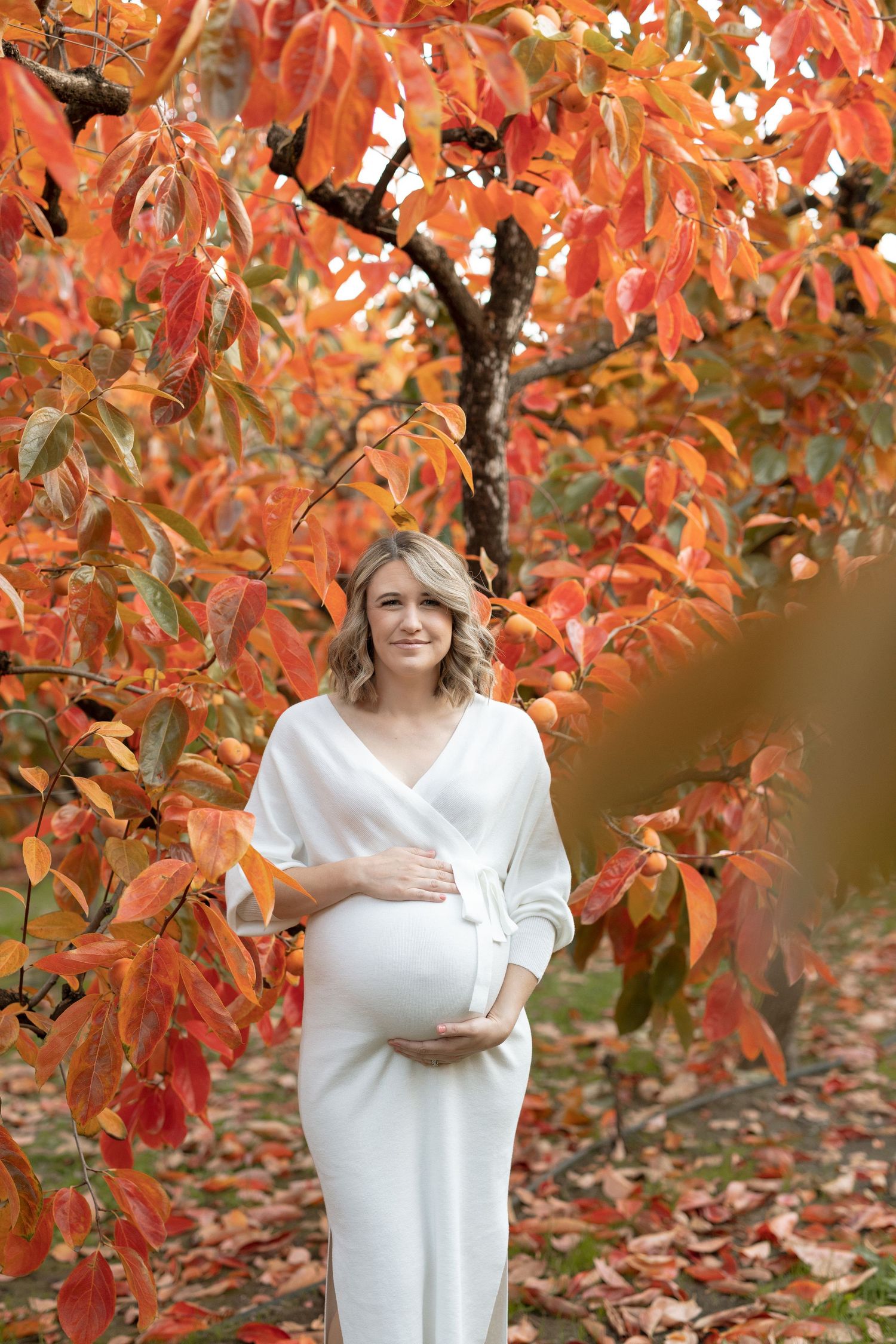 Expectant mother in white dress poses against backdrop of orange fall foliage.