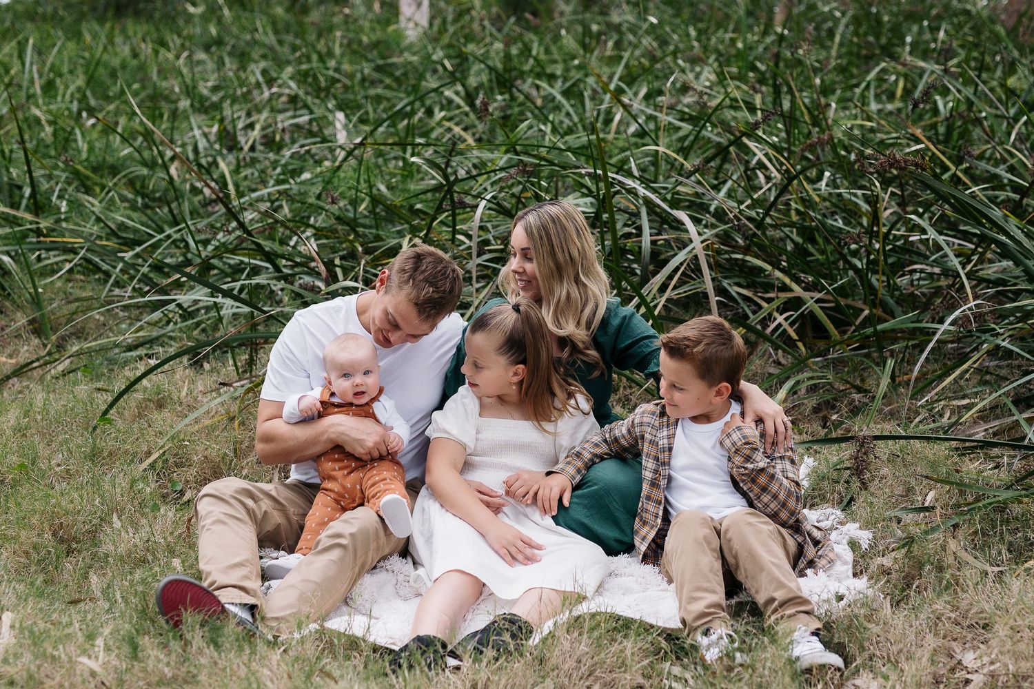 Family sitting together on grass surrounded by tall reeds during outdoor photo session.