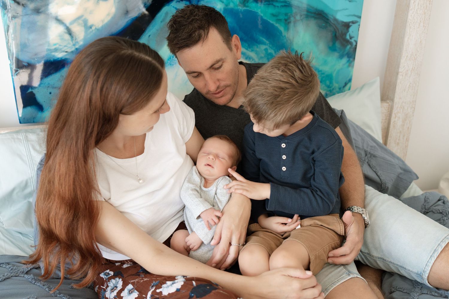 A family gathered on bed with turquoise artwork in background sharing intimate moment.