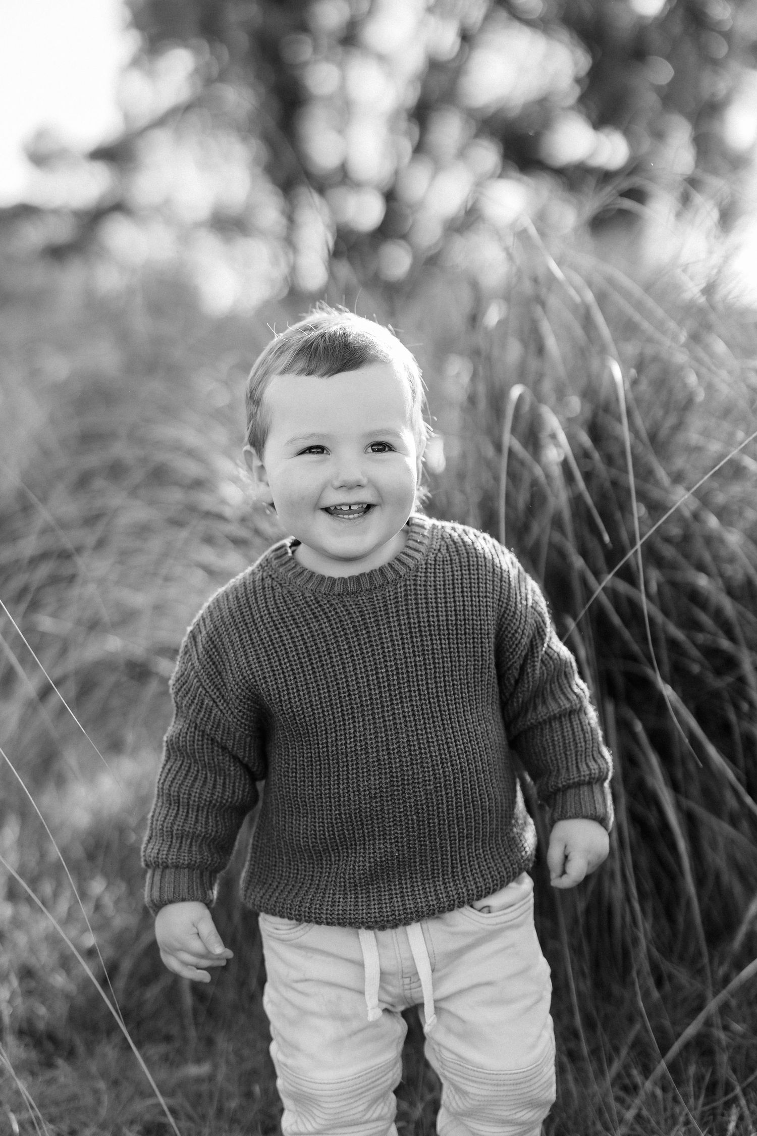 Toddler in gray sweater smiling while walking through tall grass.