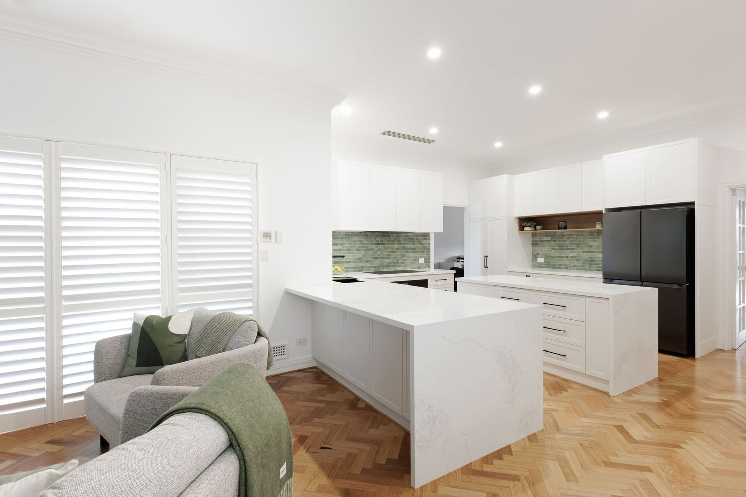 Modern white kitchen with marble island, herringbone timber floors, green subway tile backsplash, and open-plan living area.
