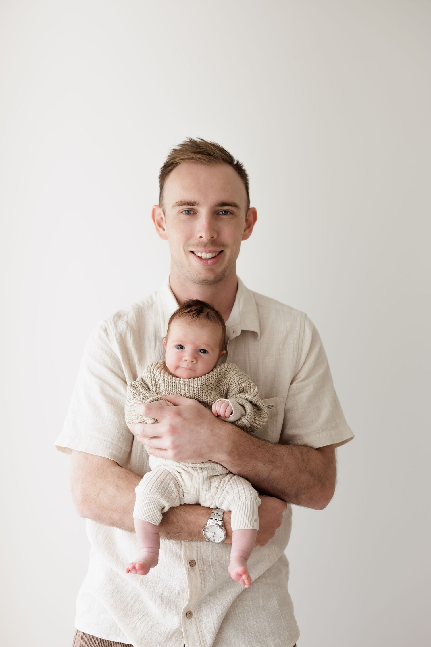 Father in beige shirt holds infant wearing knit outfit against white background.