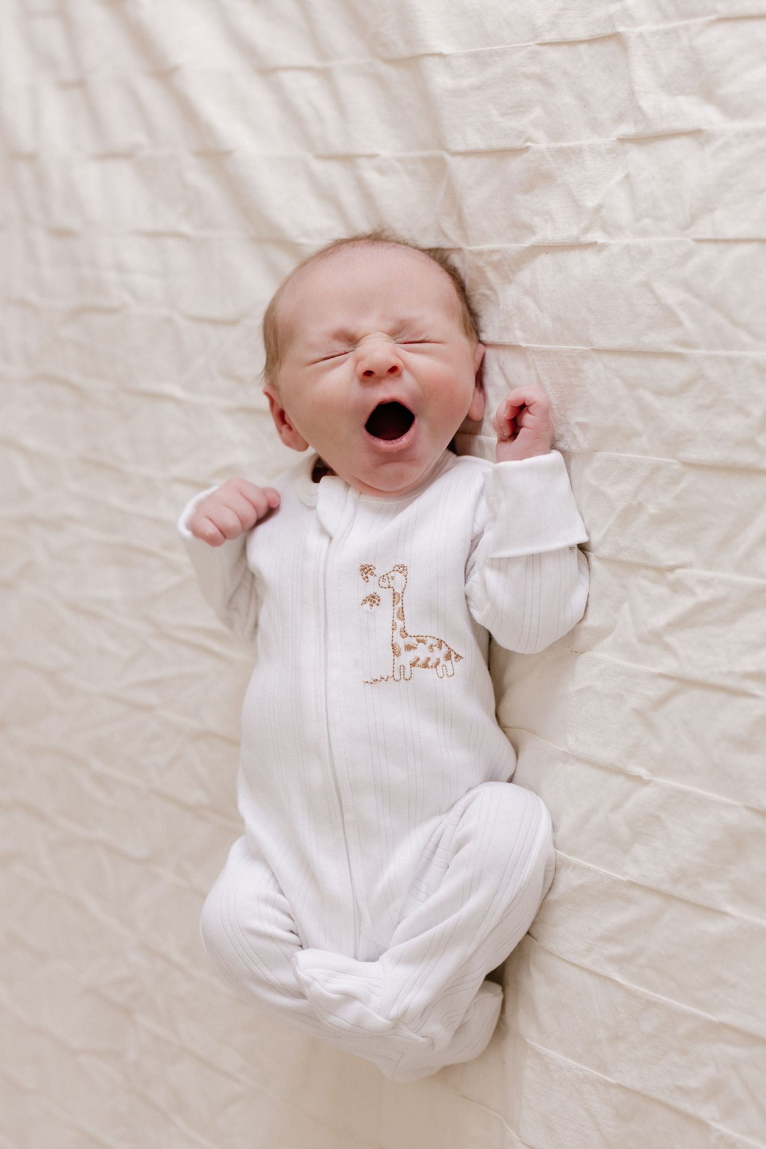 Newborn baby in white onesie lying on cream colored fabric showing different expressions.