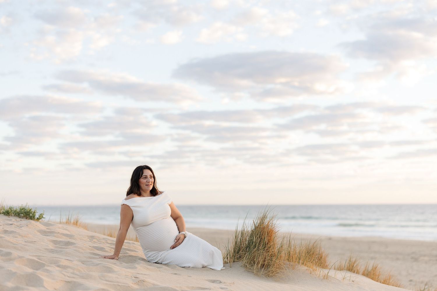 Pregnant woman in white dress sitting on sand dunes at beach during golden hour with cloudy sky.