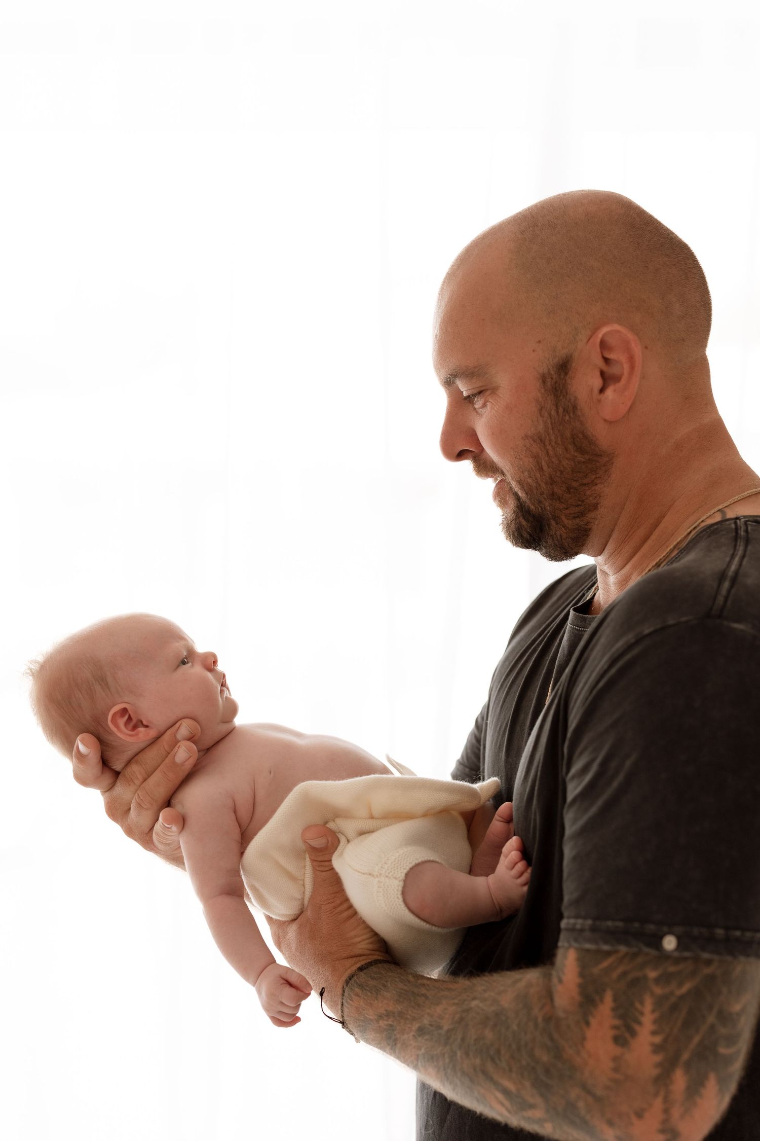 Black-clad adult with tattoos gently holding small newborn baby against white background.