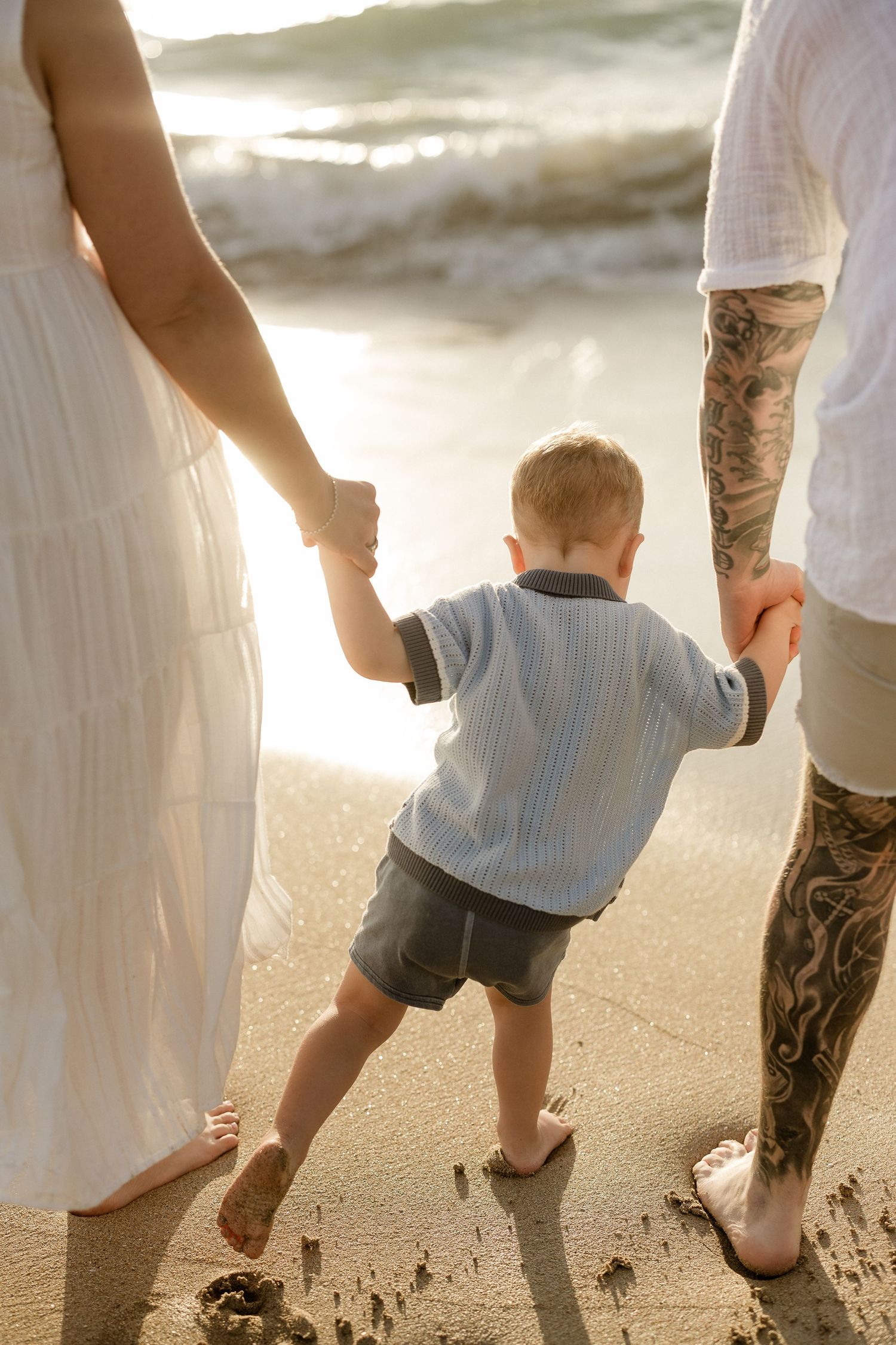 A young child holds hands with adults while standing on a sandy beach at sunset.