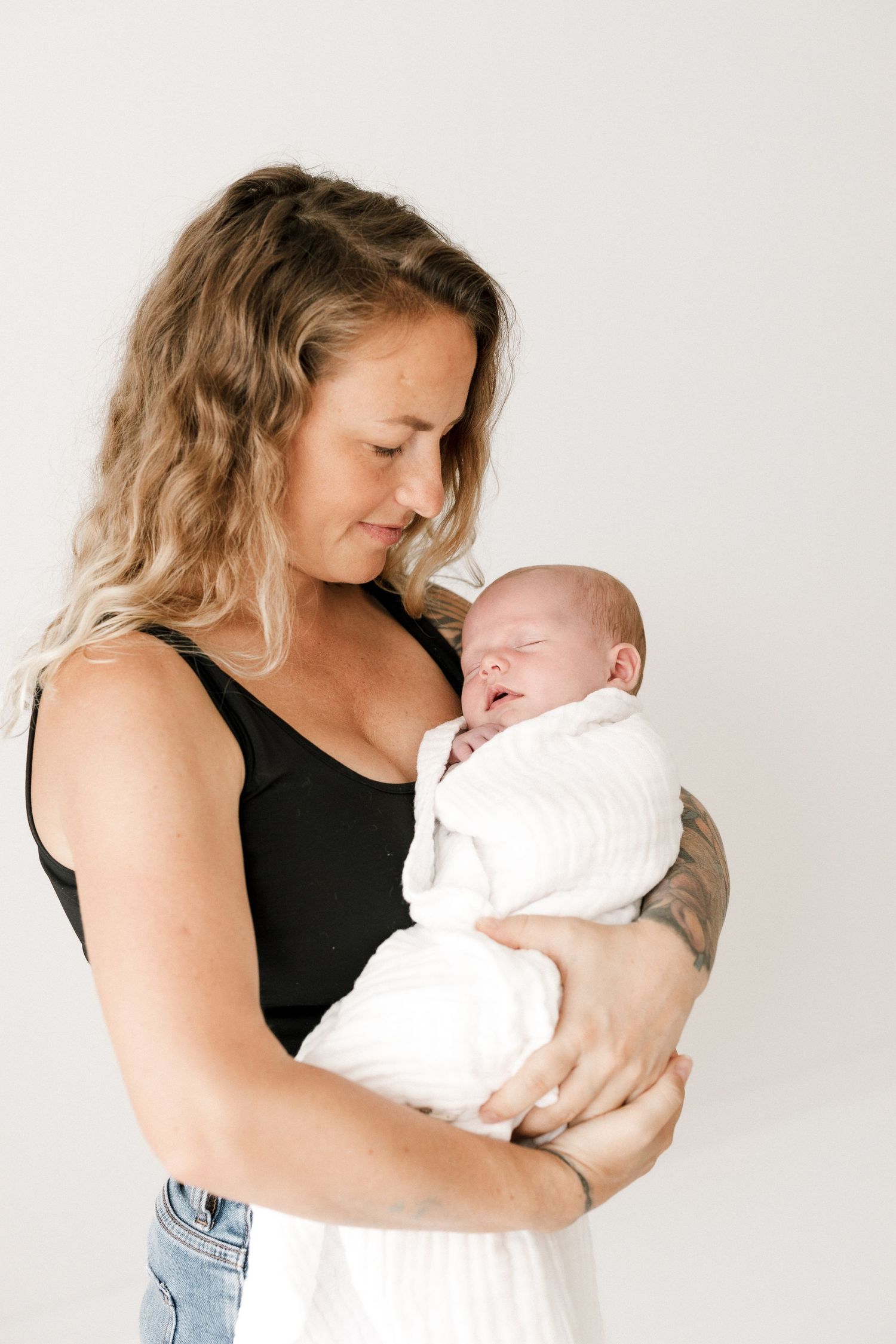 Woman in black tank top holding a sleeping newborn baby wrapped in white cloth against white background.