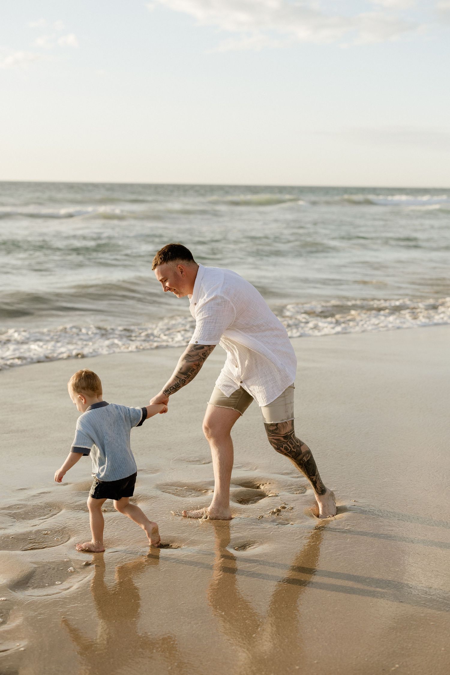 An adult and child run through the shallow ocean water on a sunny beach.