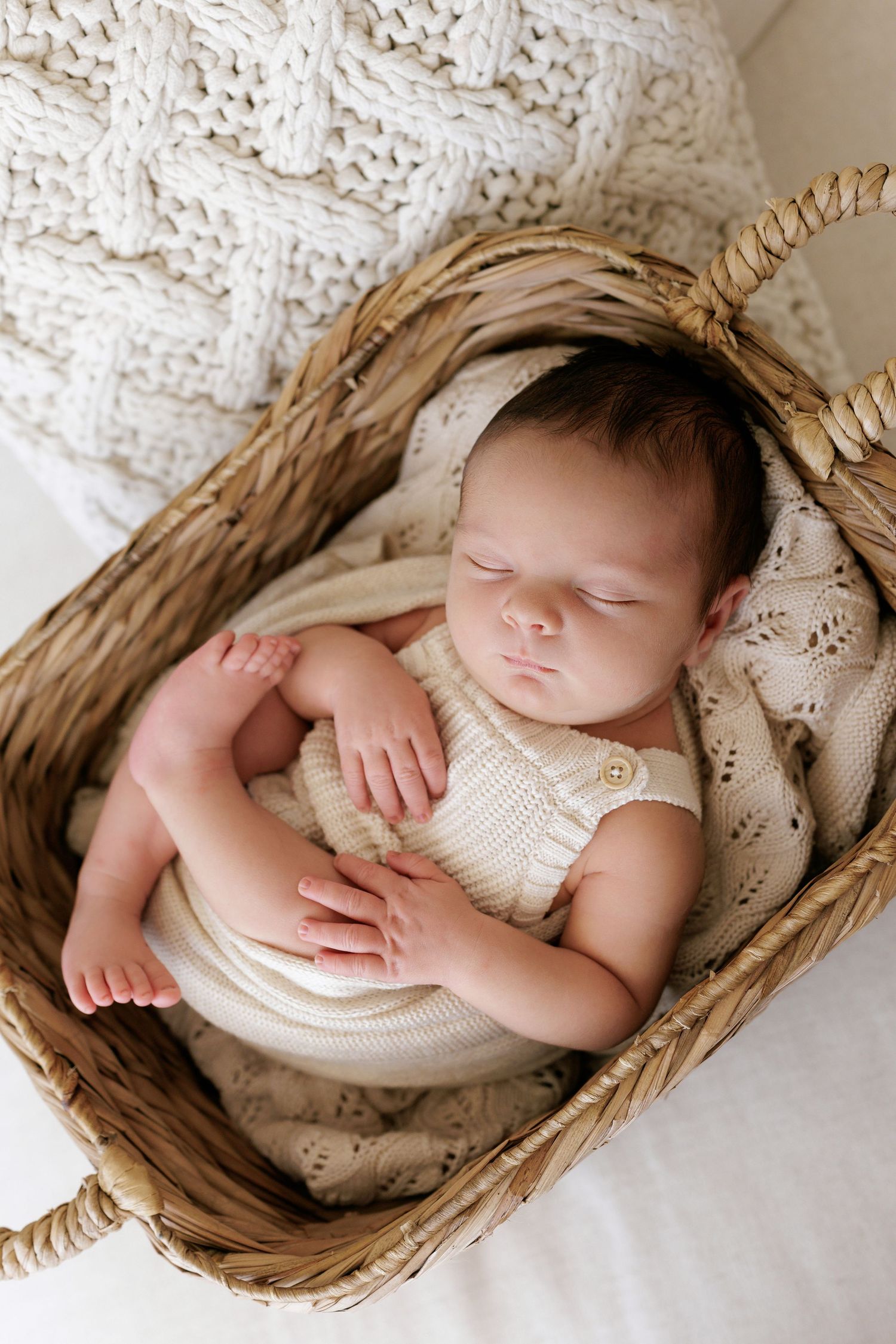 Peaceful newborn sleeping in woven basket wearing cream colored knit outfit.