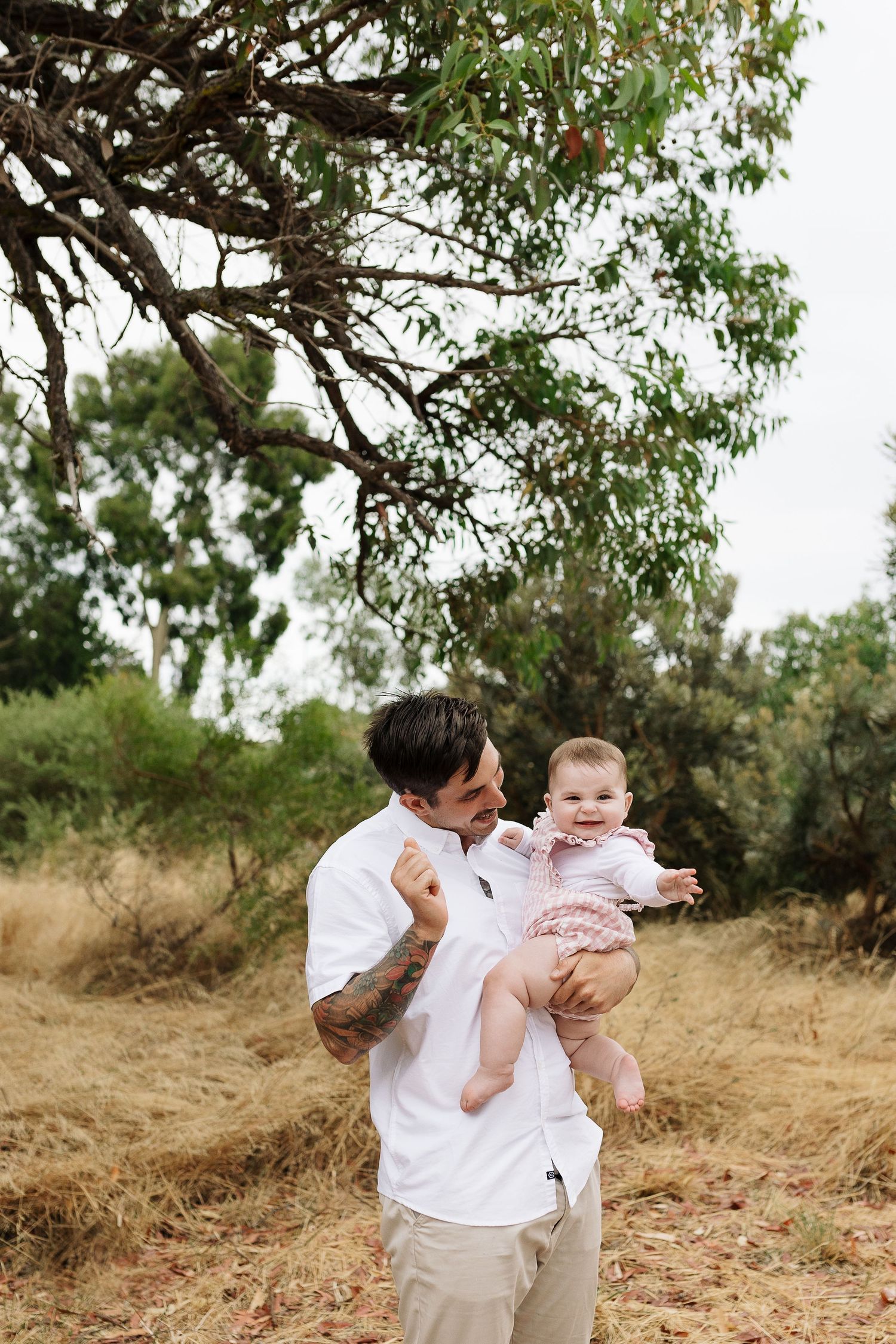 Parent holds smiling baby wearing pink outfit while standing in natural outdoor setting.