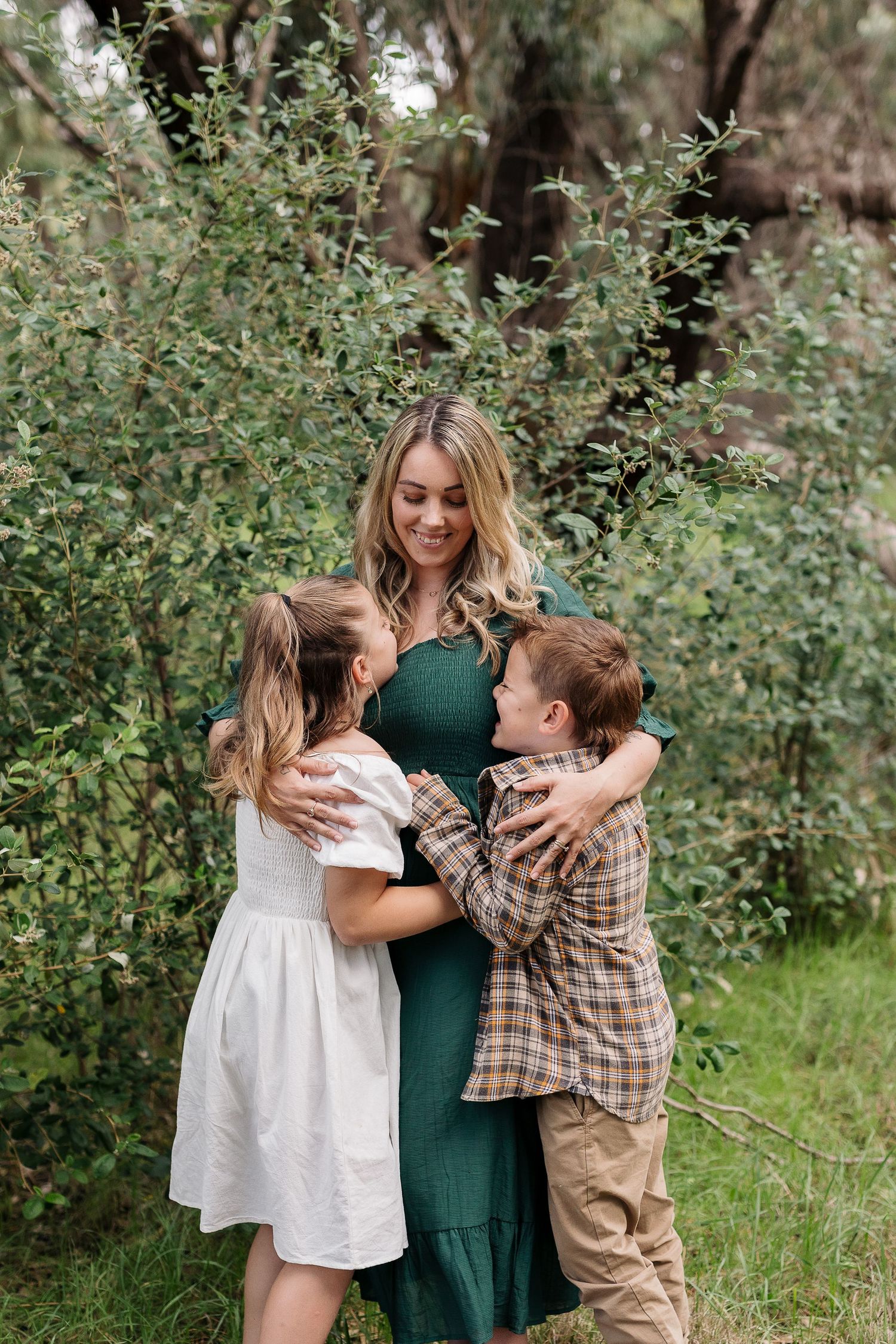 A heartwarming family moment outdoors with three people hugging and smiling among green bushes.