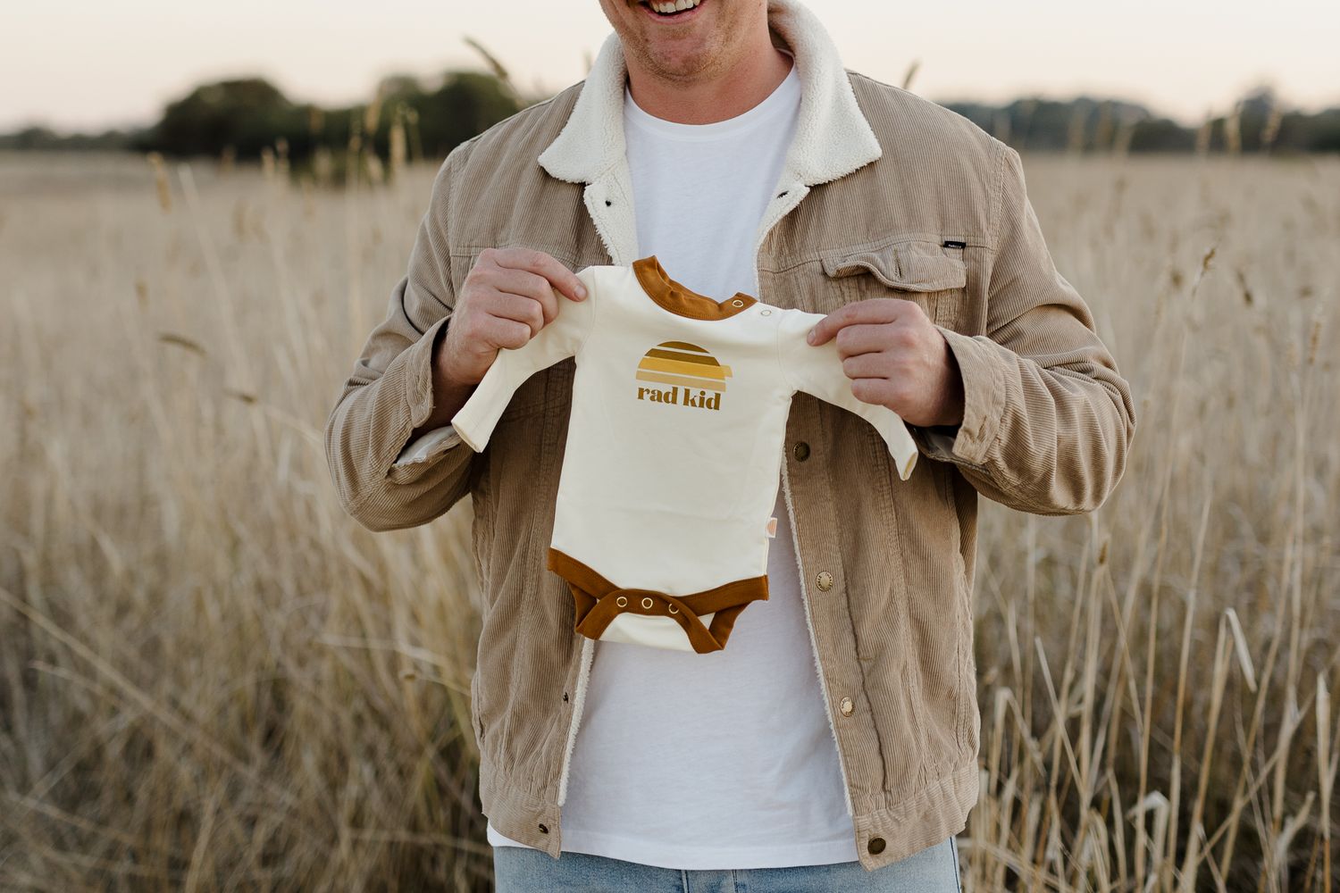 Person in tan jacket holding up small white baby onesie against field background.