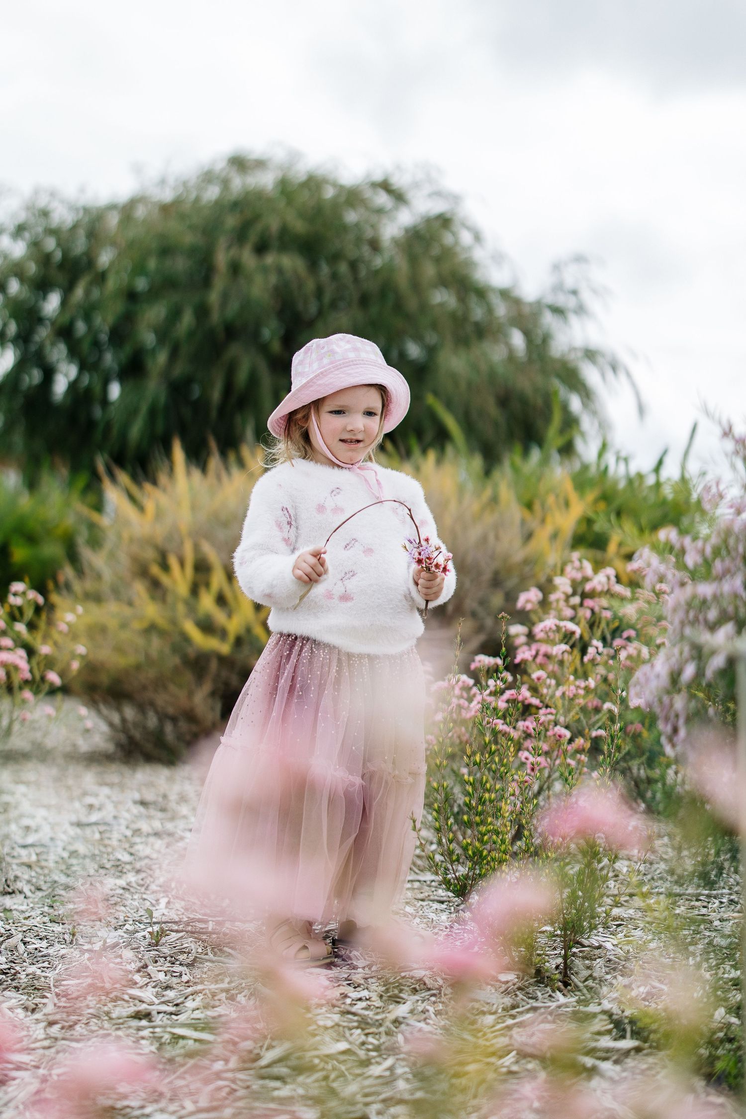 Whimsical garden portrait with pink flowers and matching outfit against cloudy sky.