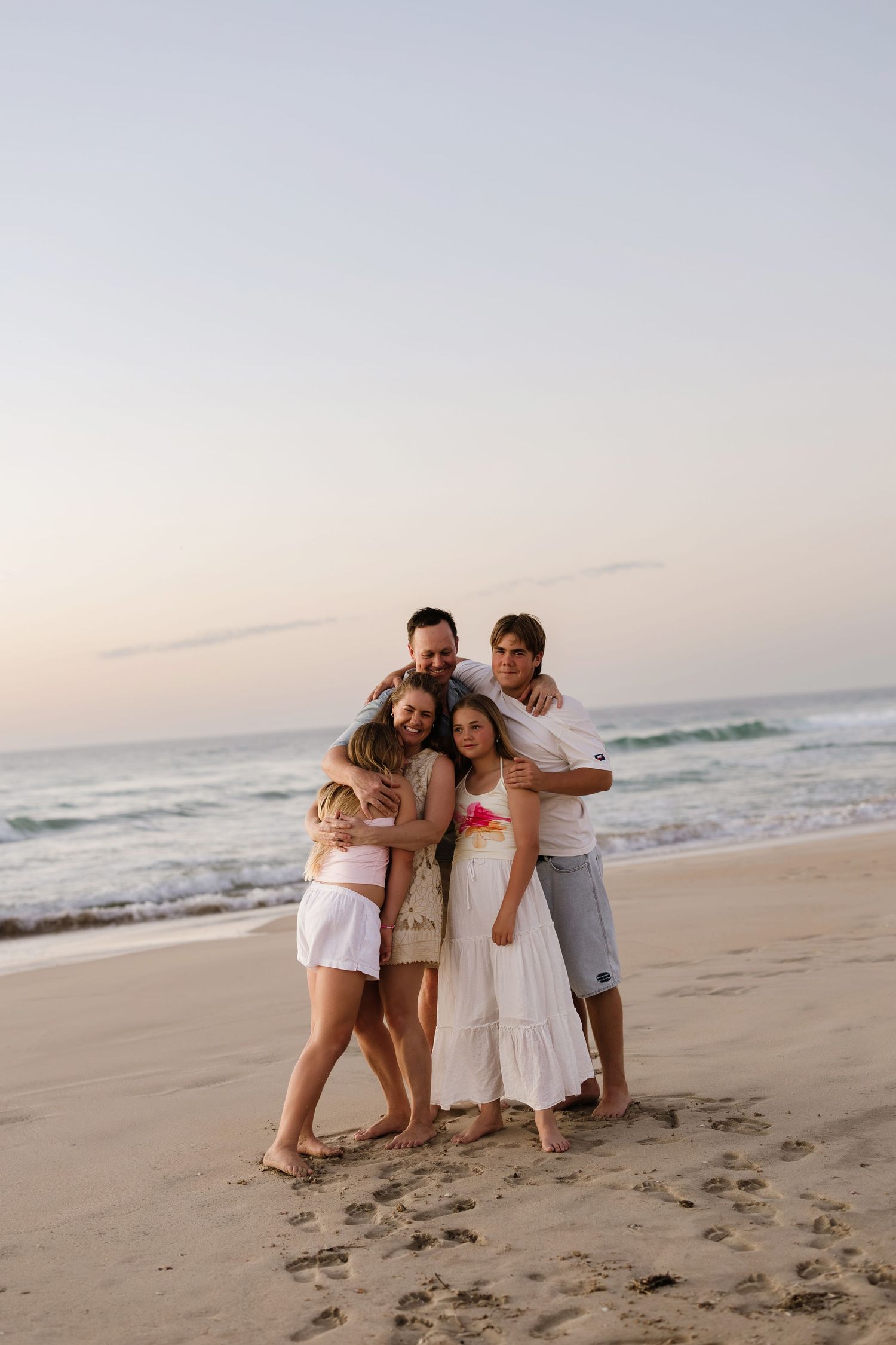 A family embraces while standing together on a beach at sunset with ocean waves in the background.